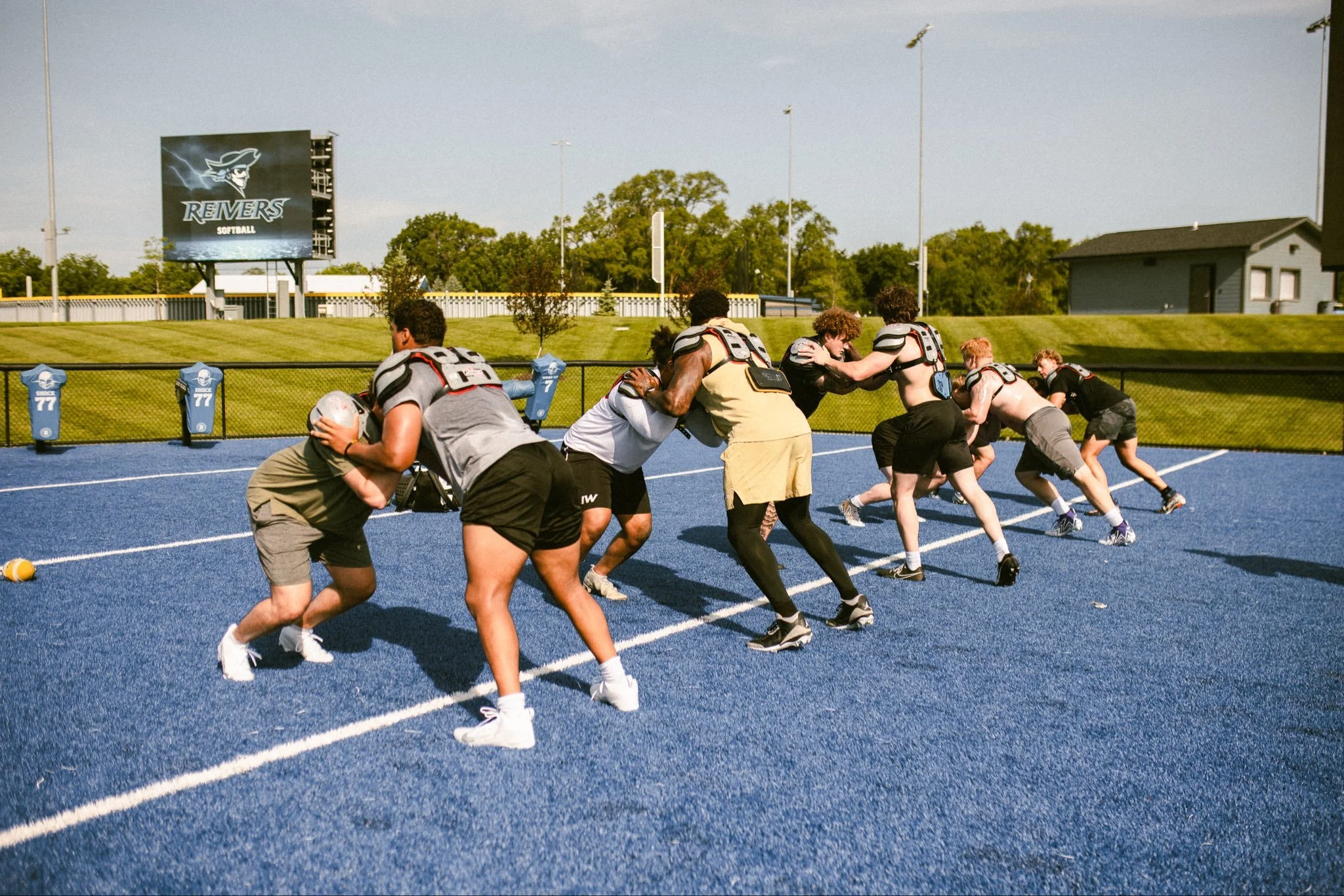 Young athletes compete in a flag football game on a blue outdoor field, with a scoreboard and trees in the background. #DLINEU Pass Rush Summit Iowa Western Football