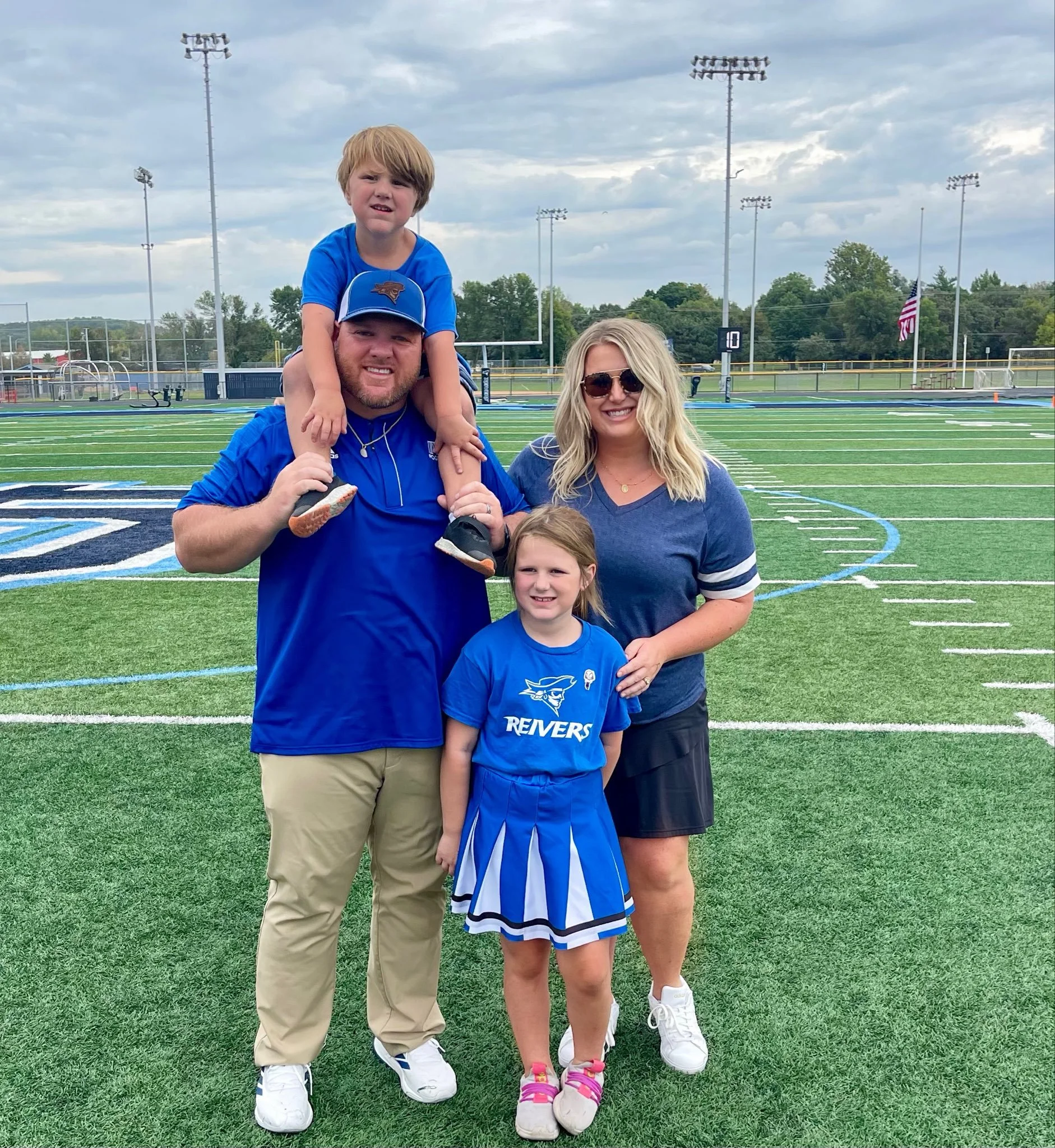 A family of four posing on a sports field, with a young boy on his father's shoulders, a woman, and a girl in front. They are wearing blue sports shirts, and the boy is wearing a blue cap. The background features a football field, stadium lights, and an American flag.