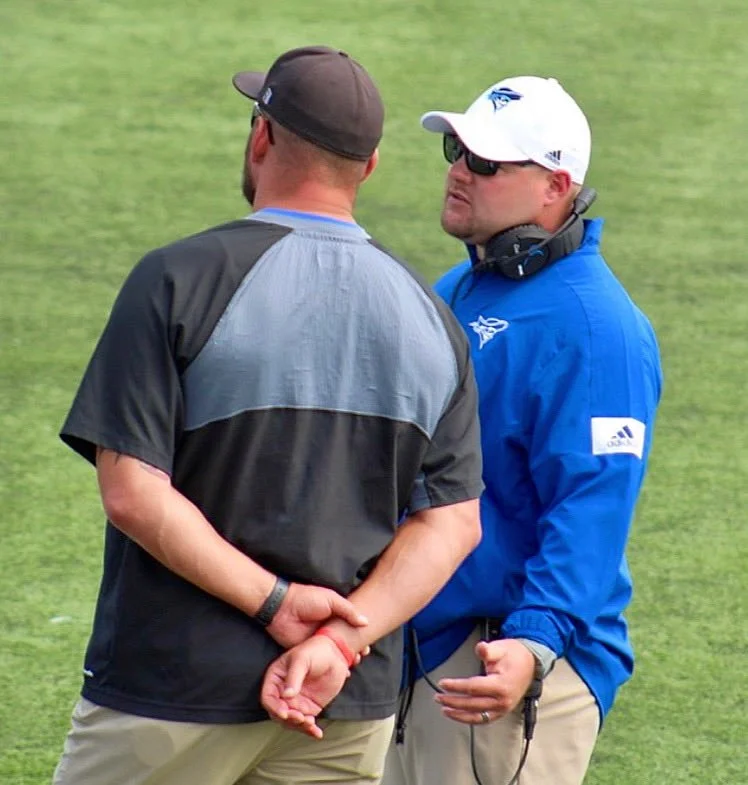 Two men having a conversation on a golf course. One is wearing a black and gray shirt, a gray cap, and khaki pants, with hands clasped behind his back. The other is wearing a blue jacket, white cap with a logo, sunglasses, and khaki pants, with a headset around his neck.