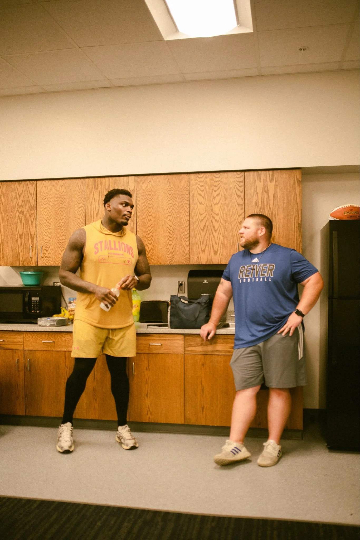 Two men standing in a room near wooden cabinets, engaged in conversation. One is wearing a yellow athletic outfit with