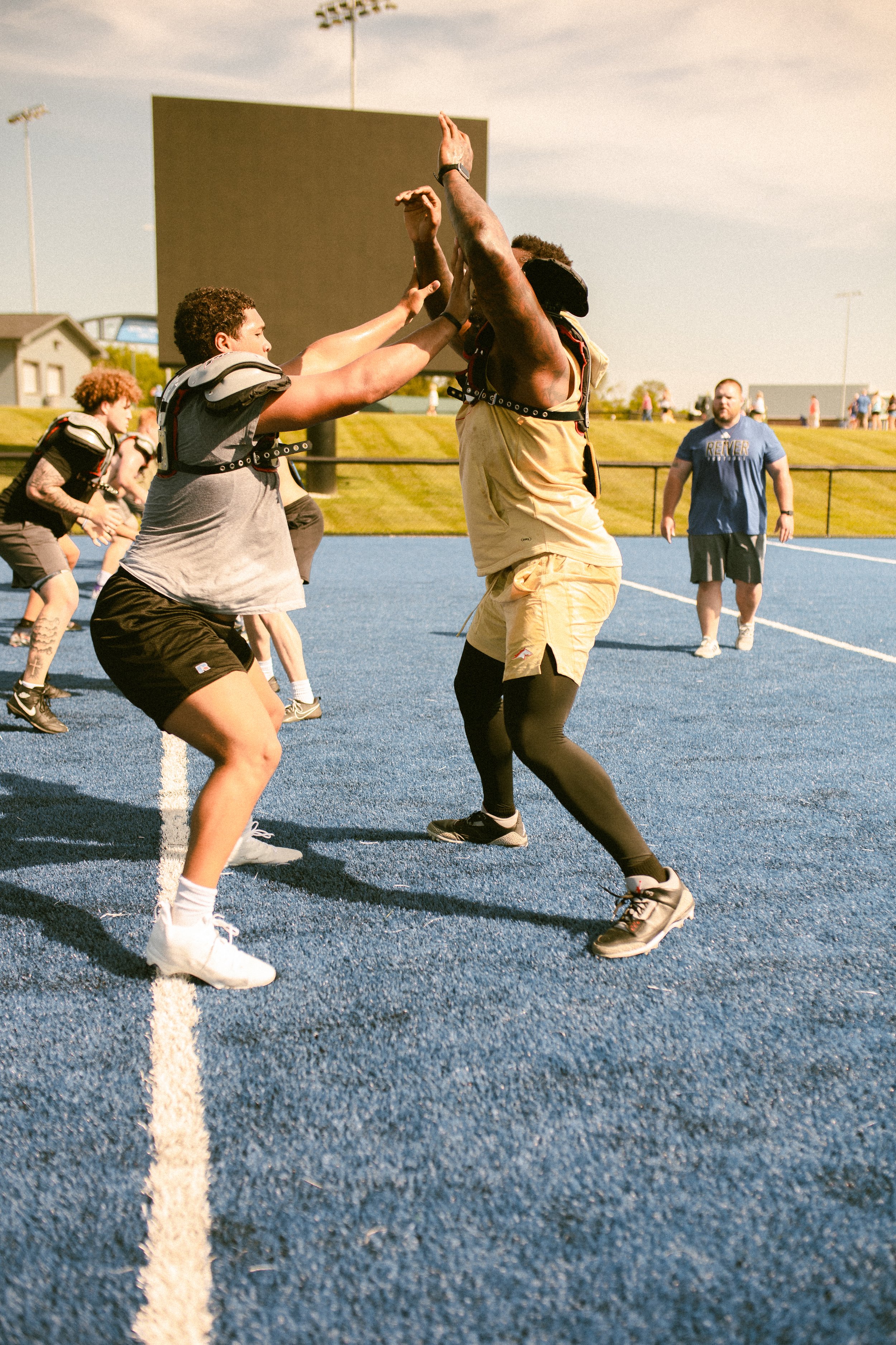 #dlineu Pass Rush Summit Football Perrion Winfreydoing drills on a blue turf field during daytime practice or game, with spectators and field lights in the background.