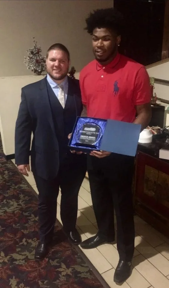 Two men standing indoors, one in a suit and the other in a red polo shirt, holding a plaque or award in a blue folder. A small decorated Christmas tree is visible in the background.