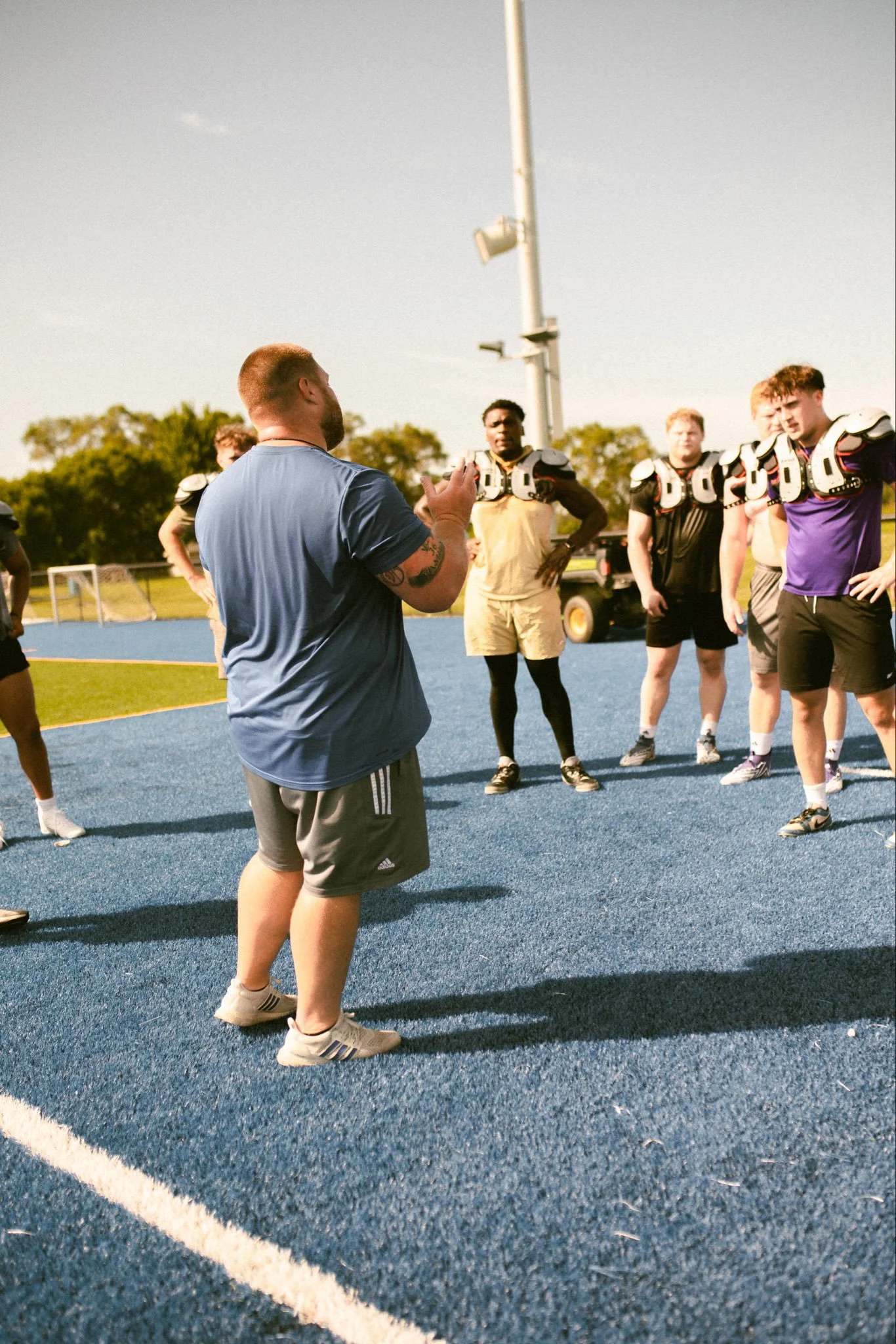 #DLineU Iowa Western Football Coach Aaron Terry giving instructions to a group of football players during practice on a blue outdoor field. The players are wearing football pads and casual athletic clothing, listening attentively.