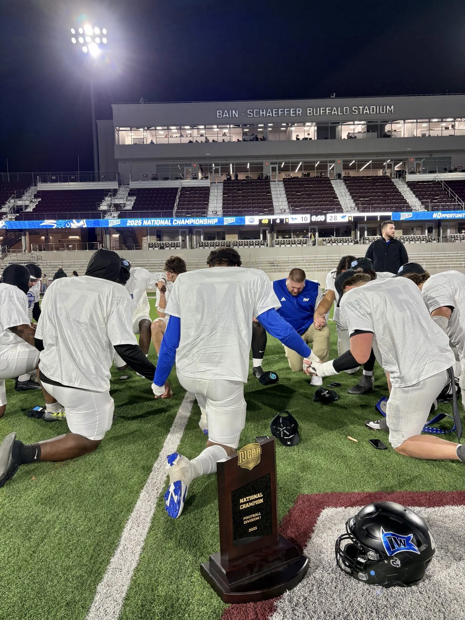 Football team kneeling on field during night game at Bain-Schaeffer Buffalo Stadium after winning national championship, with trophy and helmet visible.