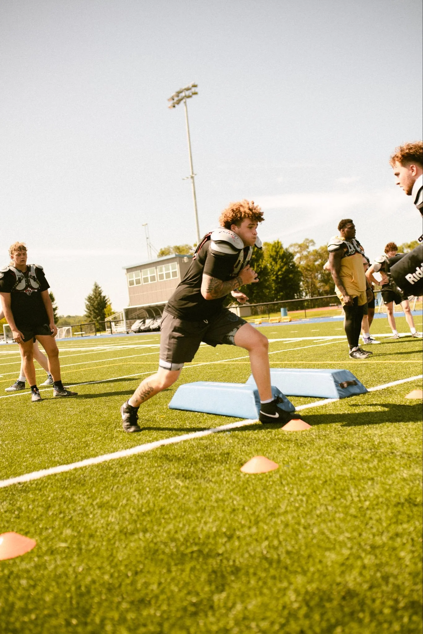 #DLINEU Pass Rush Summit Iowa Western Football Young male athlete in black sports uniform and shoulder pads running on a football field during training, with other players standing nearby and practice equipment such as cones and padding on the turf.