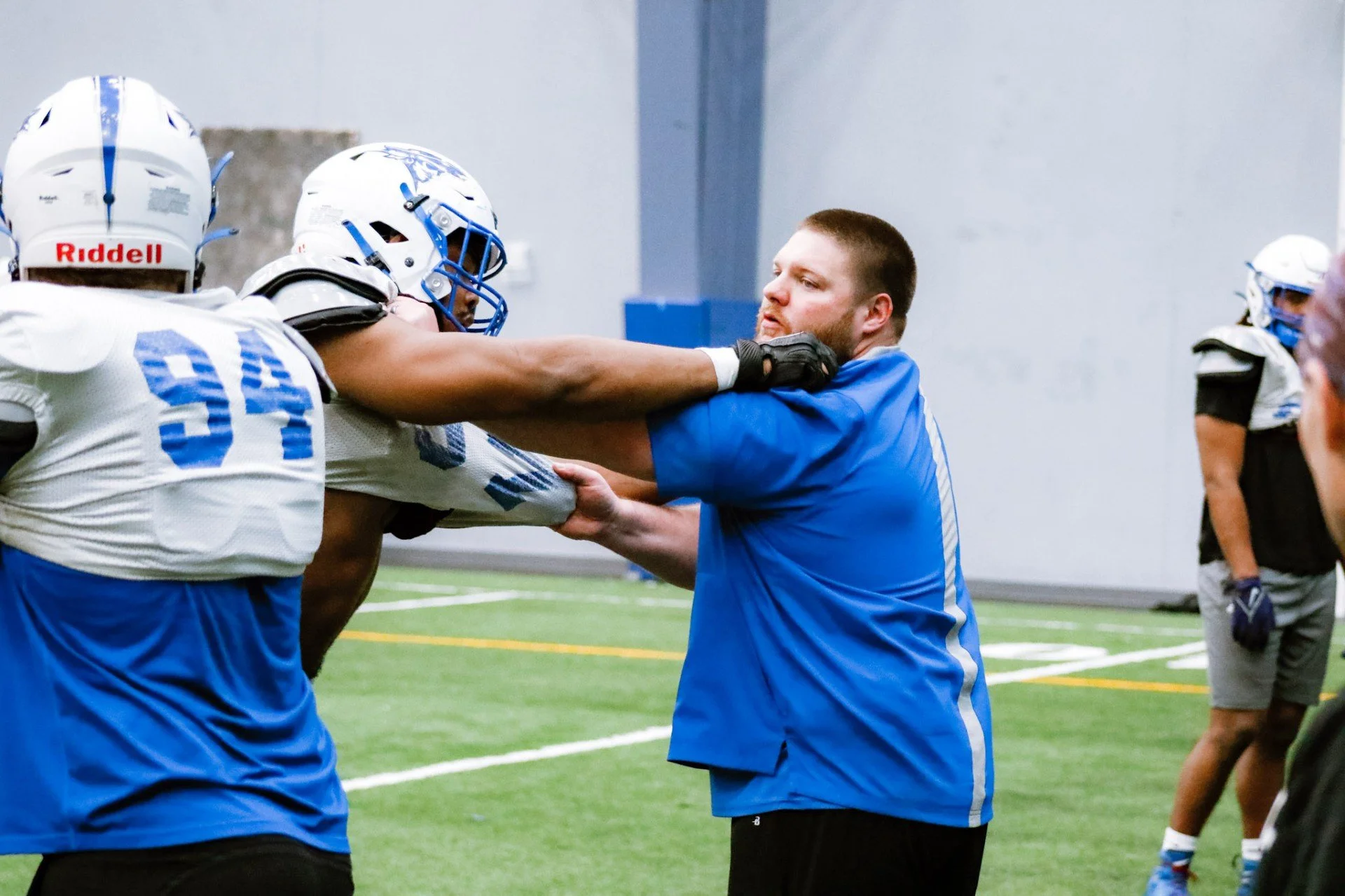 An indoor football practice where a coach or trainer is confronting or separating two players during a drill or game. The players are in full gear, including helmets, and are involved in a physical interaction.