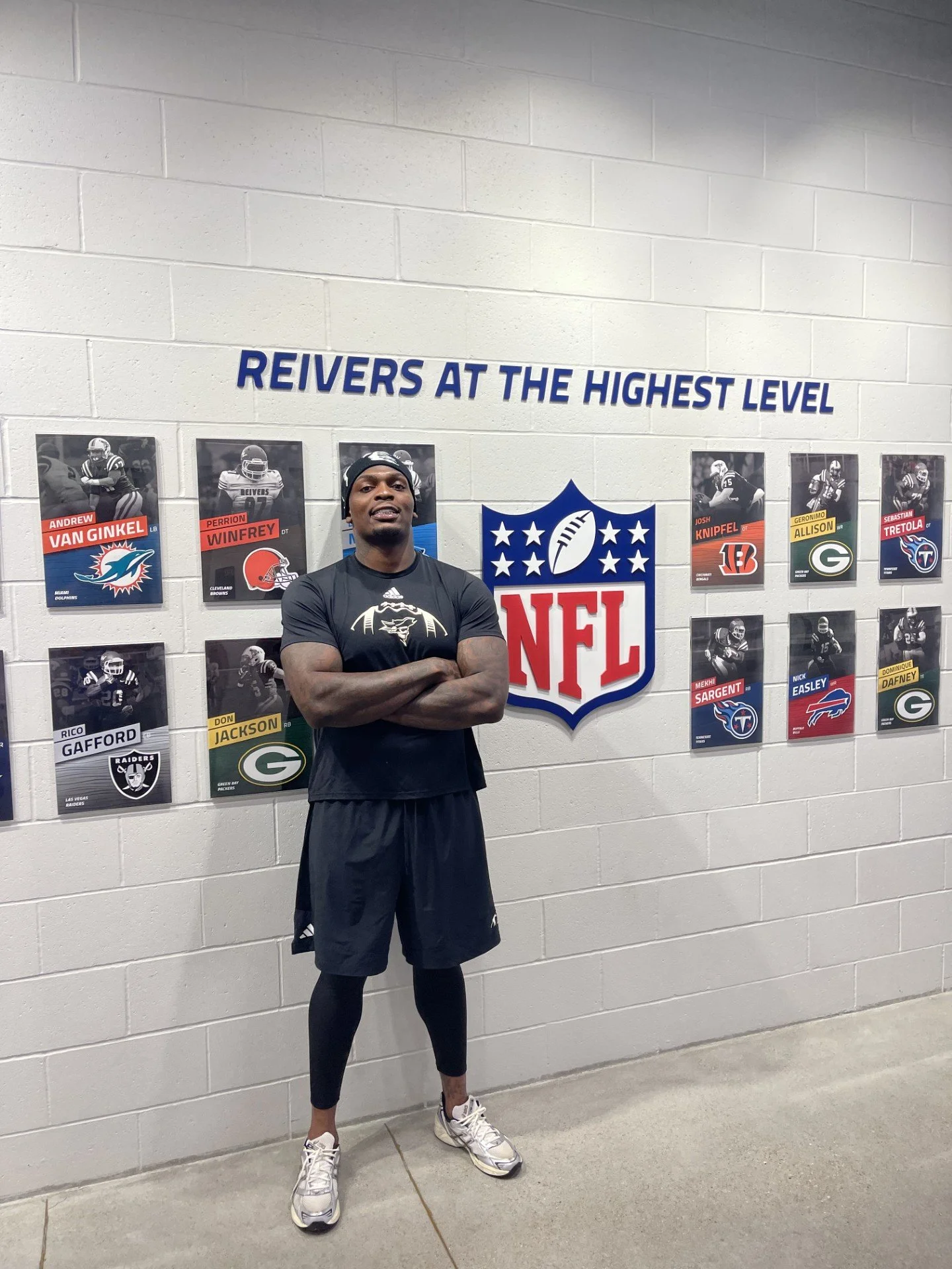 Perrion Winfrey standing with crossed arms in front of a wall displaying NFL player cards and a large NFL logo. The wall has the phrase "REIVERS AT THE HIGHEST LEVEL" written above the cards. #DLineU Iowa Western Football Player