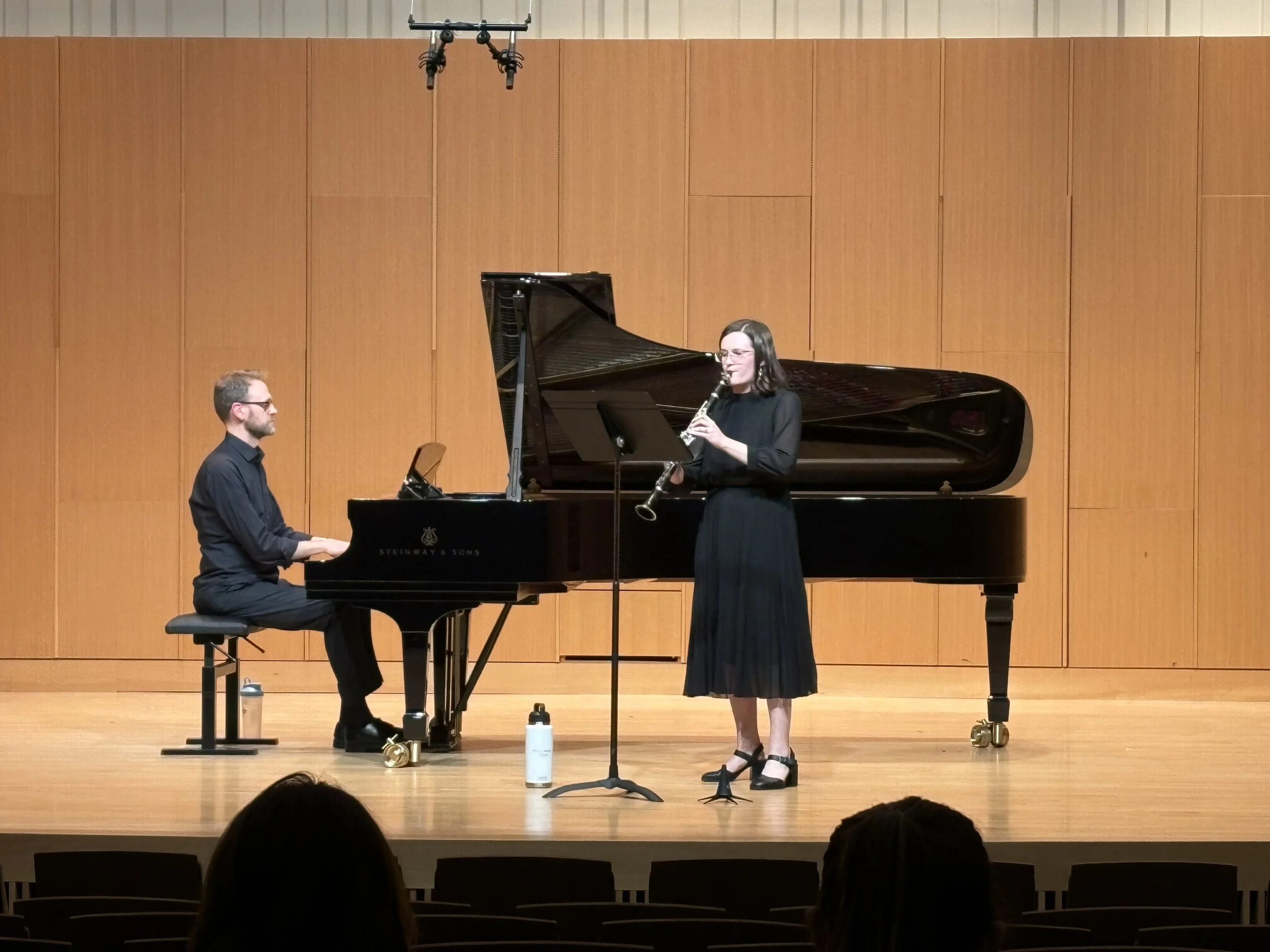 A man and a woman perform music together on stage; the man plays a grand piano while the woman plays a clarinet, both dressed formally, in a wooden hall with a large wooden wall behind them.