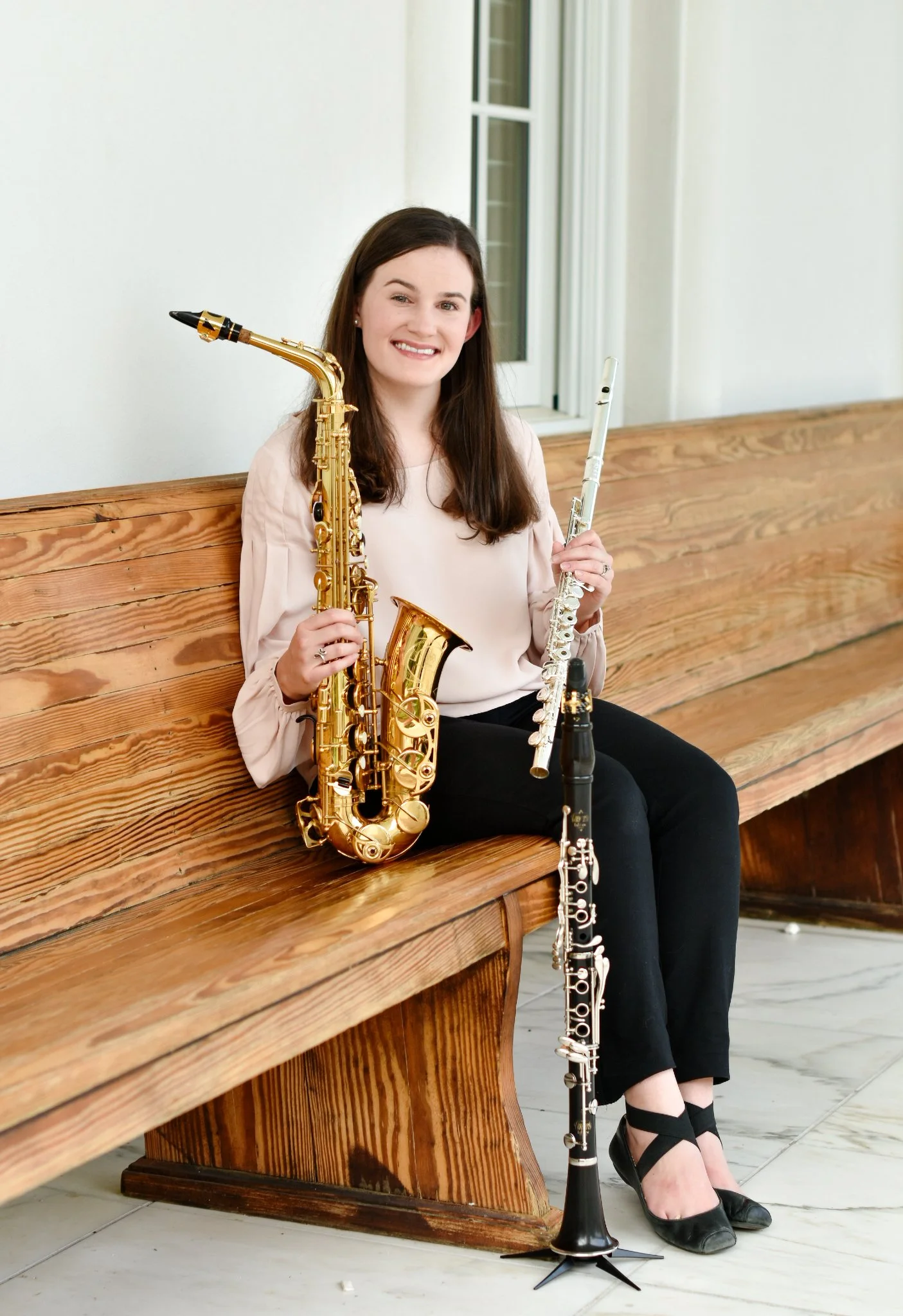 A young woman sits on a wooden bench holding a gold saxophone, with a silver flute resting on her lap, and a black clarinet on the floor beside her. She is smiling, wearing a light pink blouse, black pants, and black heels.