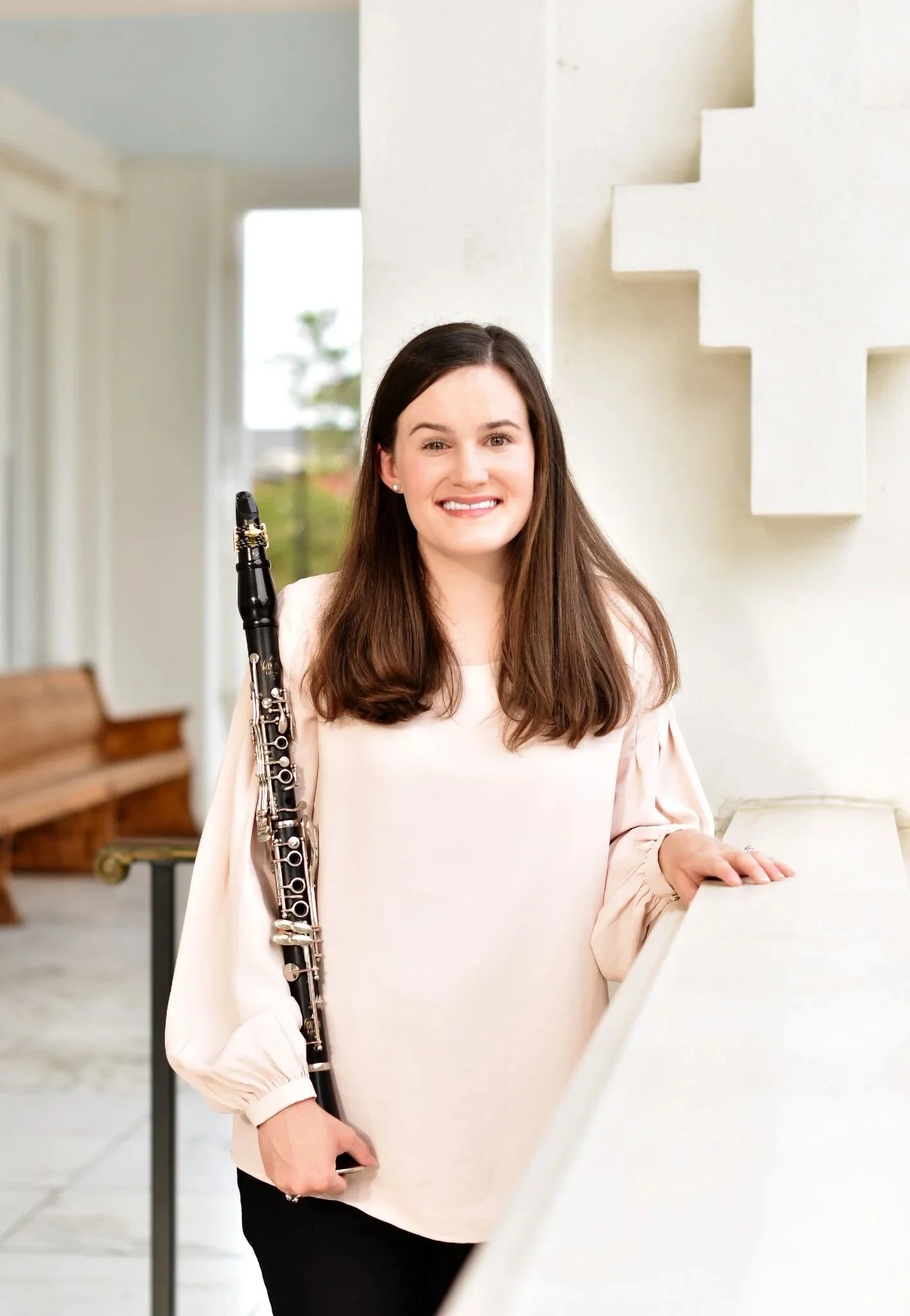 A woman with long brown hair holding a clarinet in a bright outdoor space with white walls and a staircase.