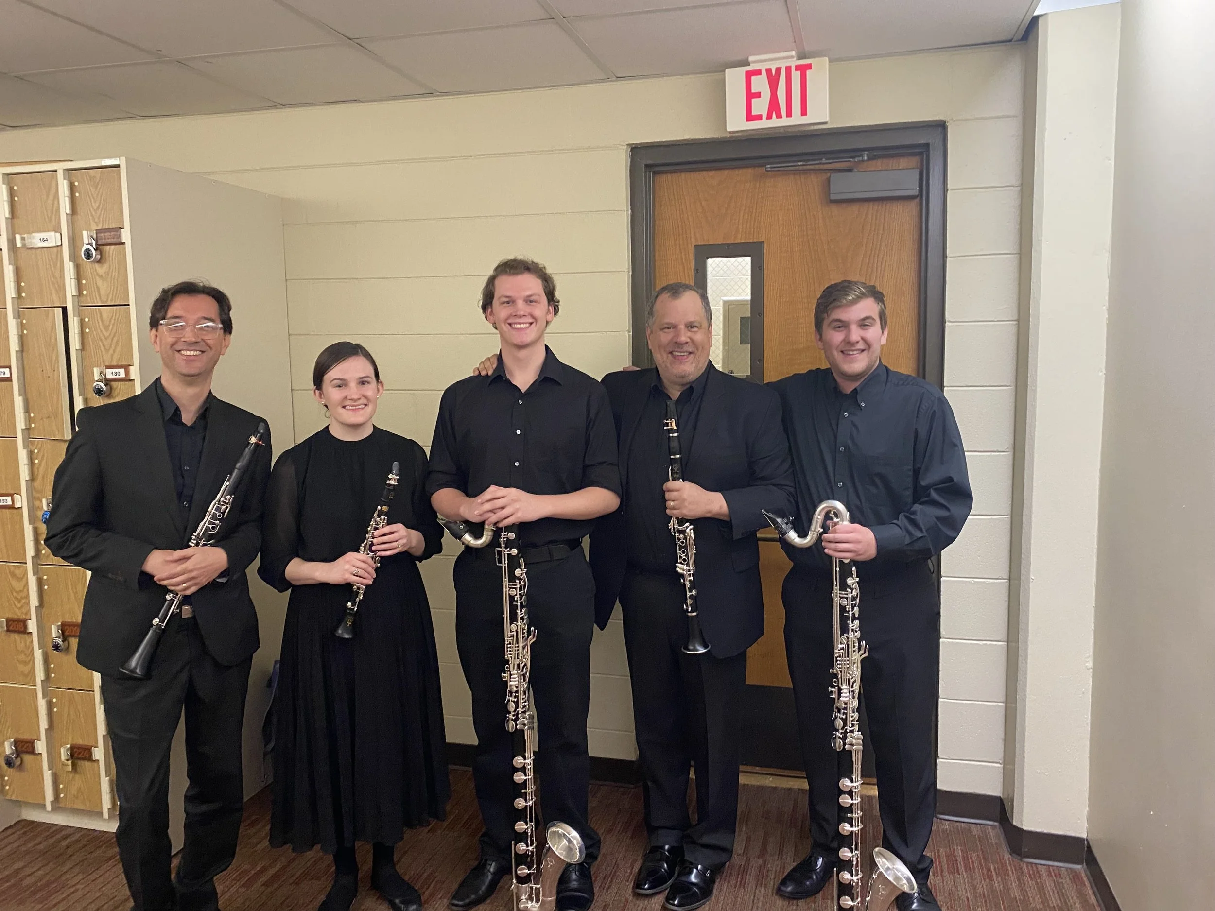 Five musicians dressed in black suits are standing together, each holding a clarinet, and smiling in front of a wall with a door and an exit sign.
