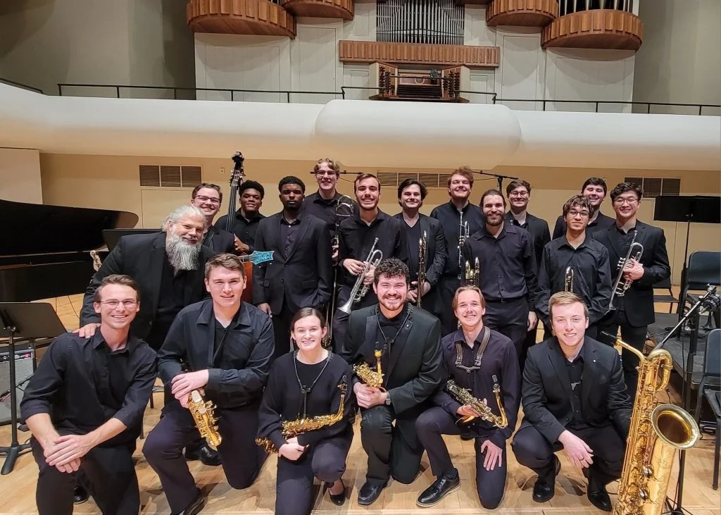 Group of young musicians with their bandleader in a concert hall, holding saxophones and trumpets, after a performance.