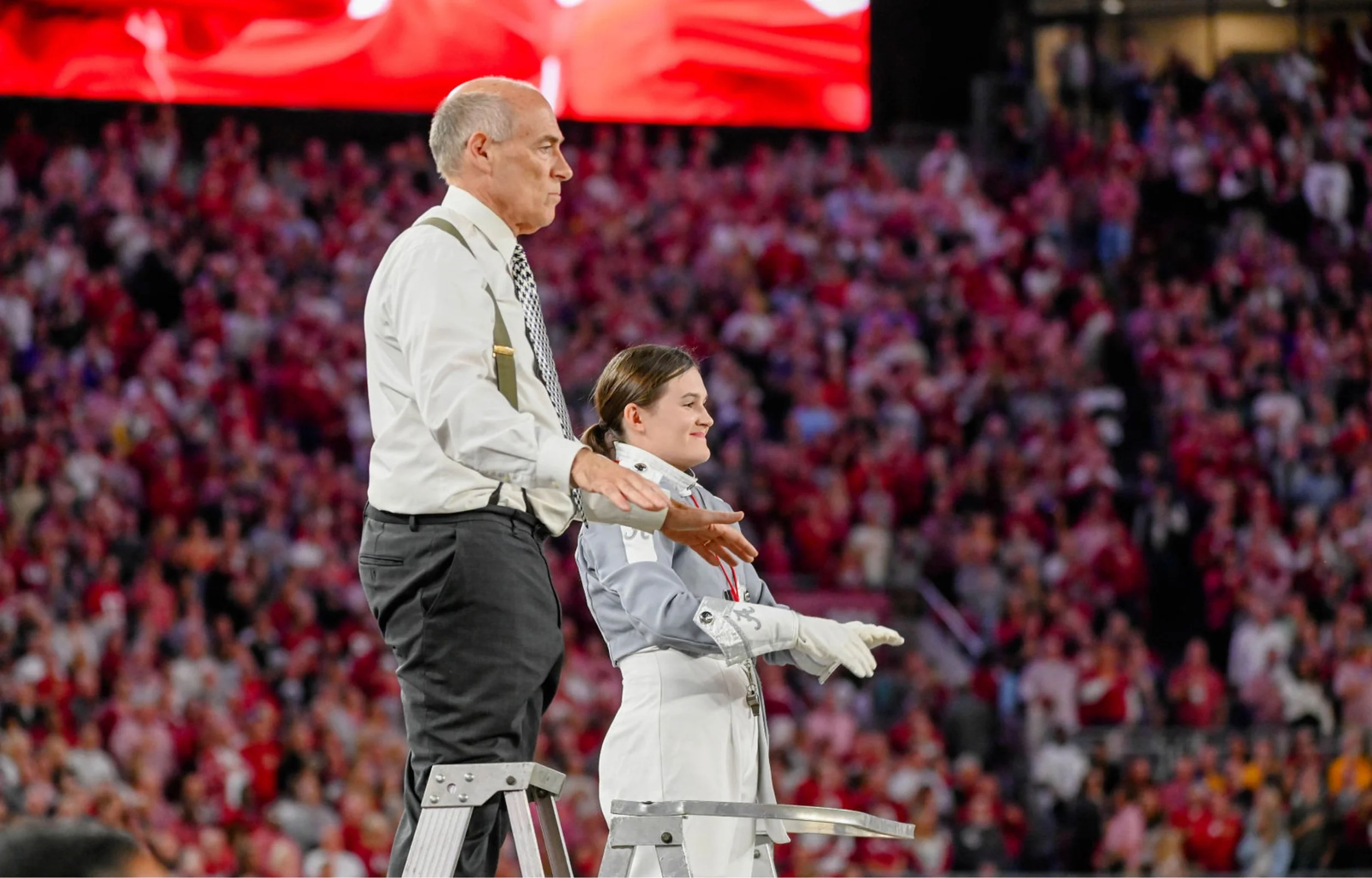 An older man with short gray hair, dressed in a white shirt, tie, and suspenders, stands next to a young woman in a white marching band uniform, both standing on a ladder at a sporting event with a large crowd in the background.