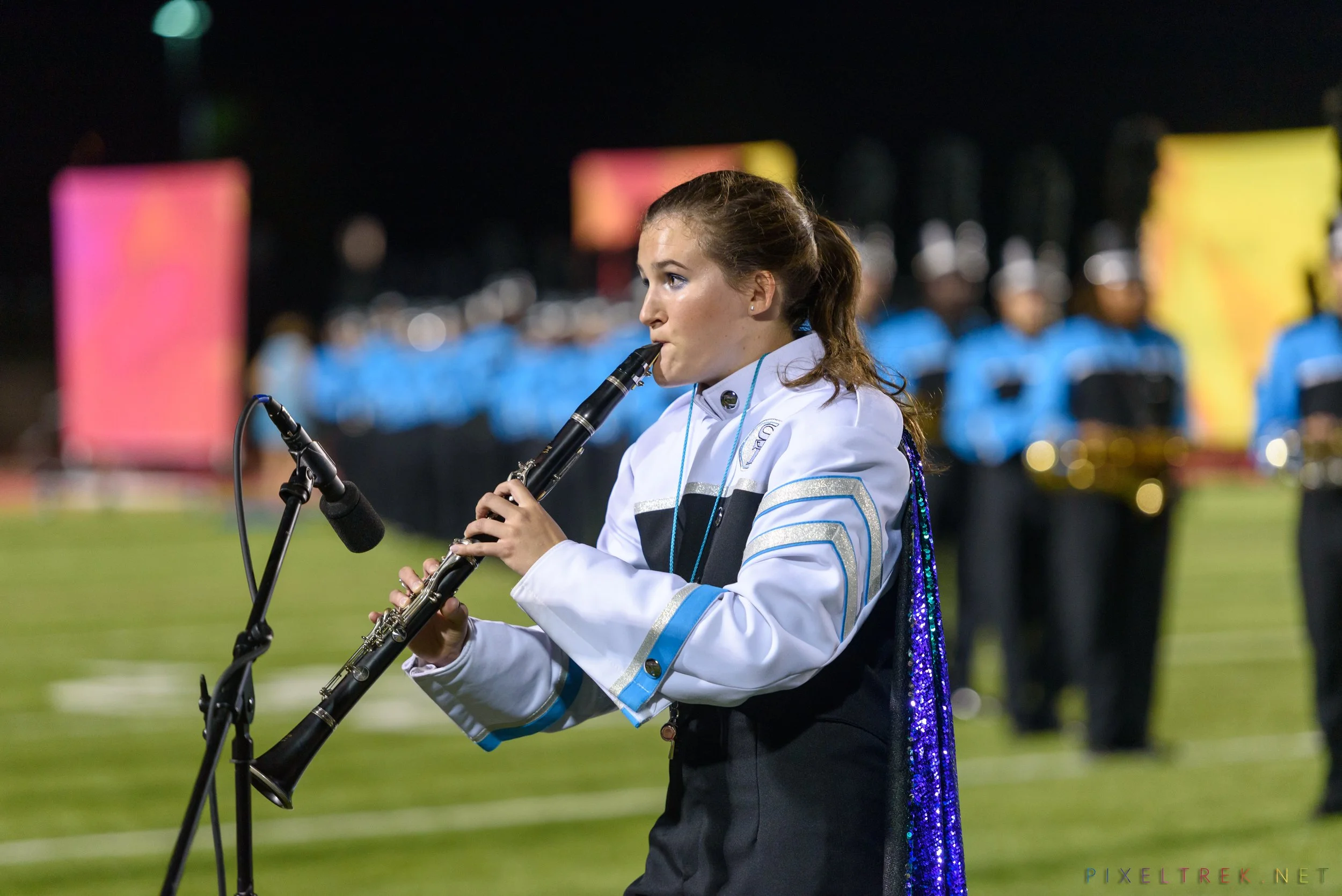 A young woman in a marching band uniform playing a clarinet on a football field at night, with others in the background.