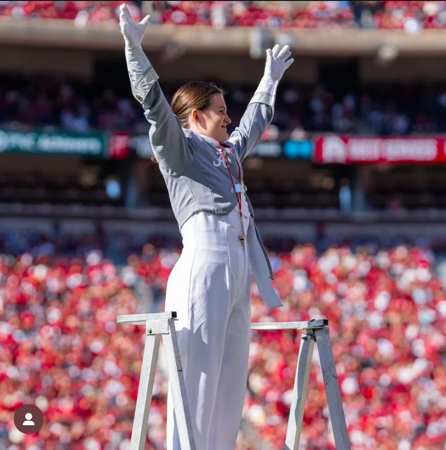 An Olympic athlete on a winner's podium with arms raised in victory, wearing a silver jacket and white pants at a stadium filled with spectators.