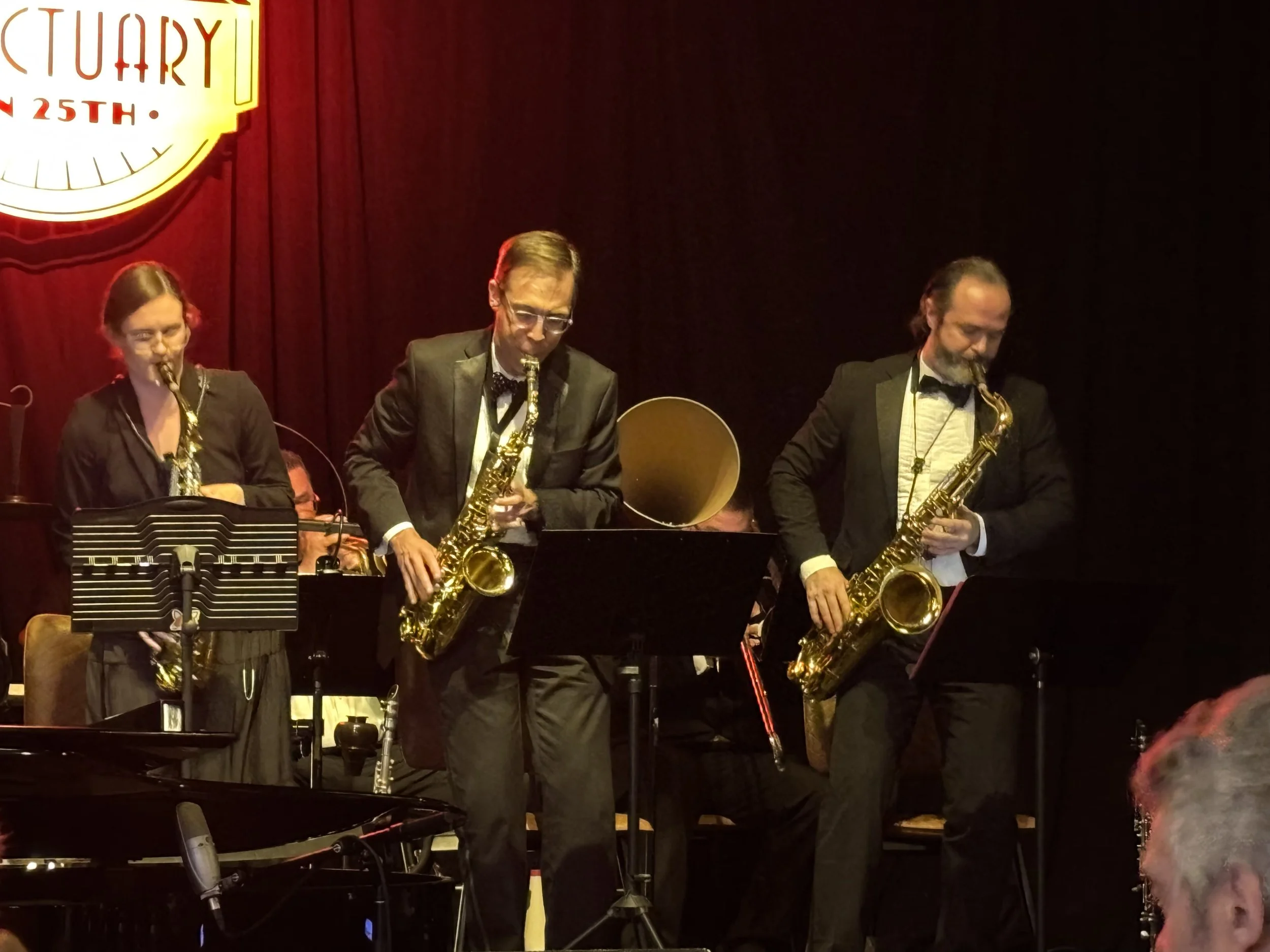 Jazz band performing on stage at a venue with a red curtain backdrop and a sign that reads 'Jazz at the 25th'. The band members are playing various instruments, including saxophones, and dressed in formal black suits with bow ties.
