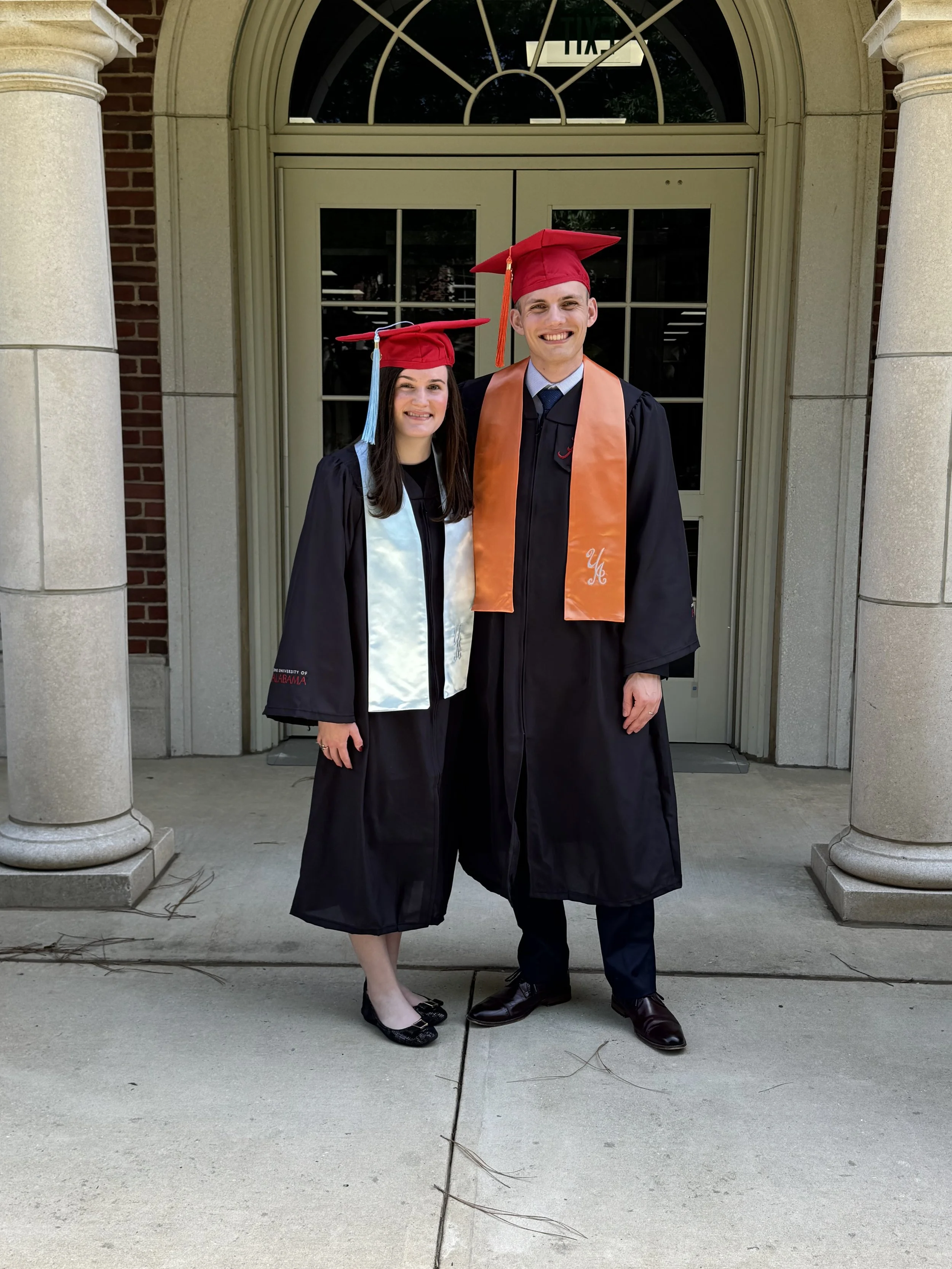 A young woman and a young man in graduation gowns and caps, standing in front of a building with columns, smiling for a photo.