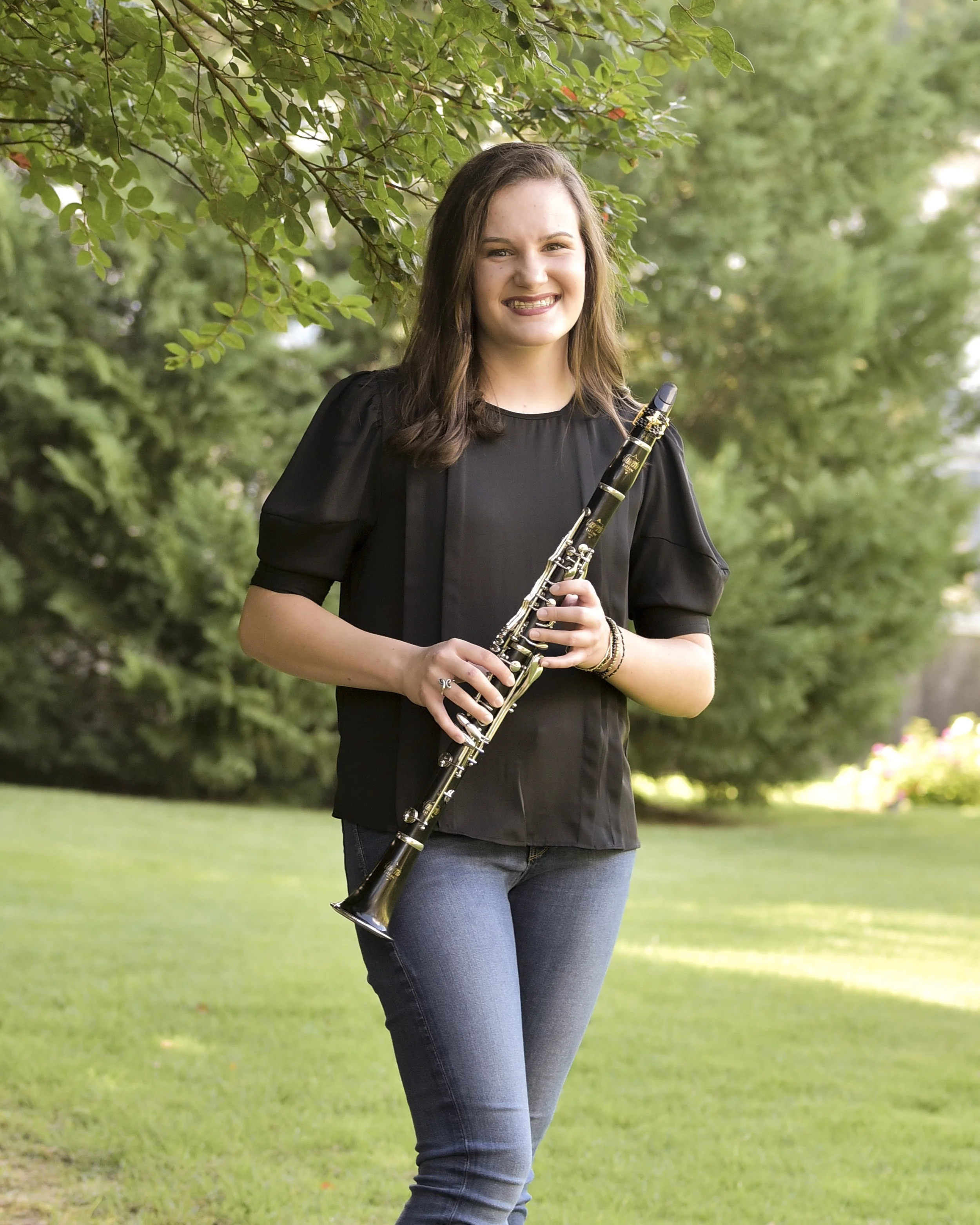 Young woman smiling and holding a clarinet outdoors, standing on grass with green trees in the background.