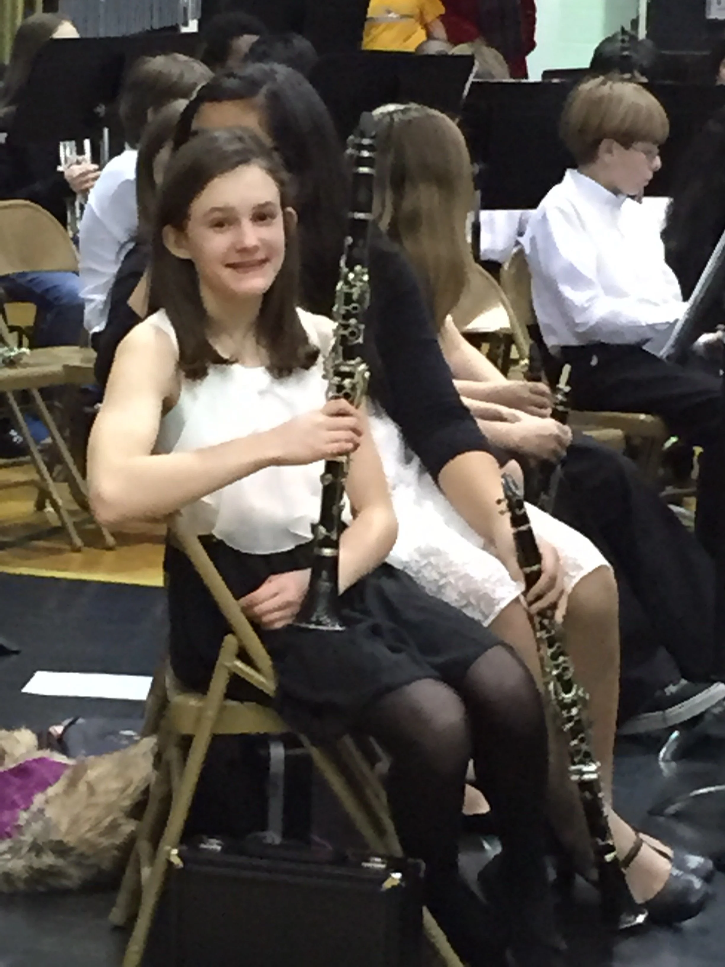 Young girl smiling and holding a clarinet, sitting in a chair surrounded by other children, some playing instruments, in a music rehearsal or classroom setting.