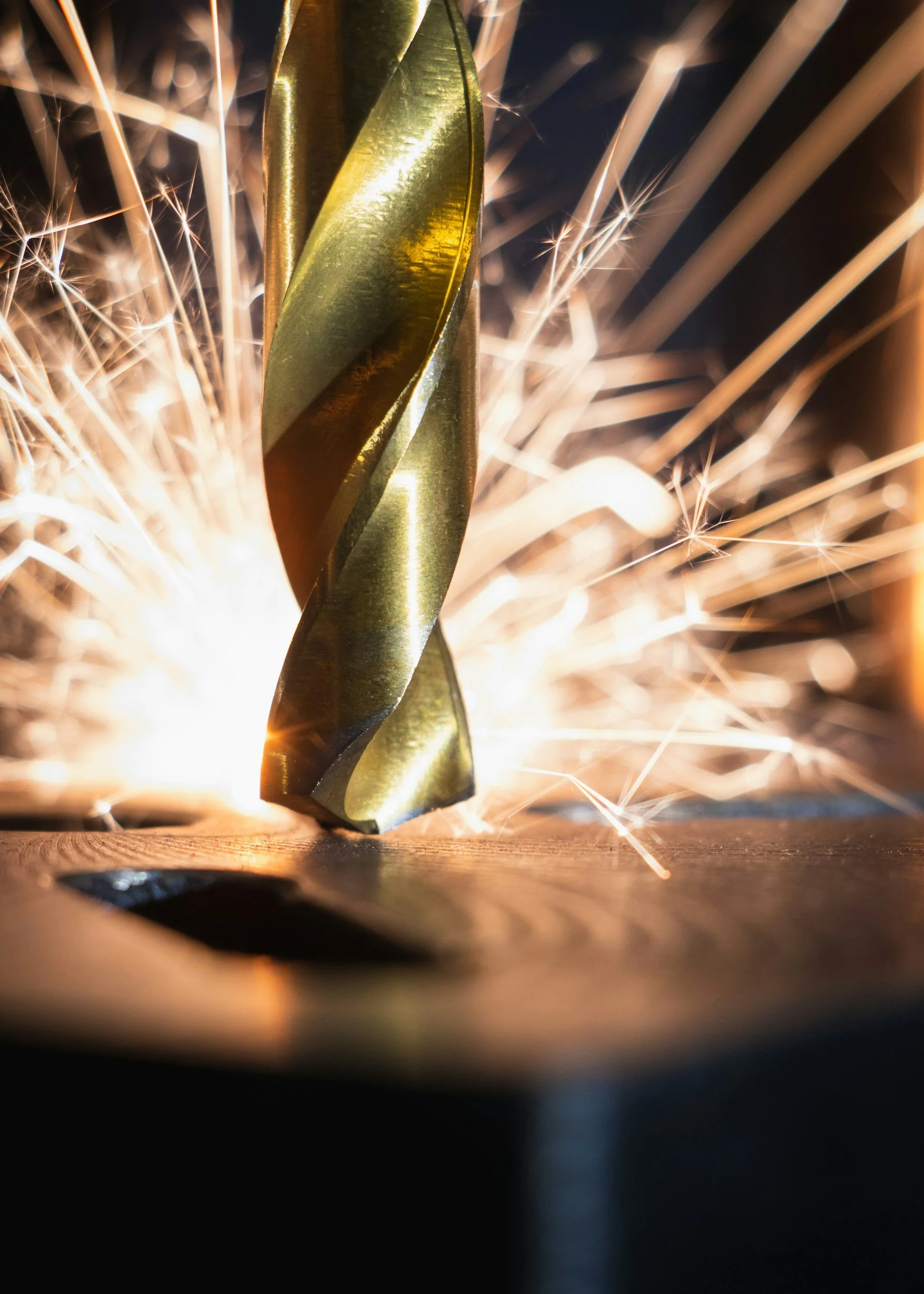 Close-up of a gold drill bit with sparks flying around as it cuts into a piece of metal.
