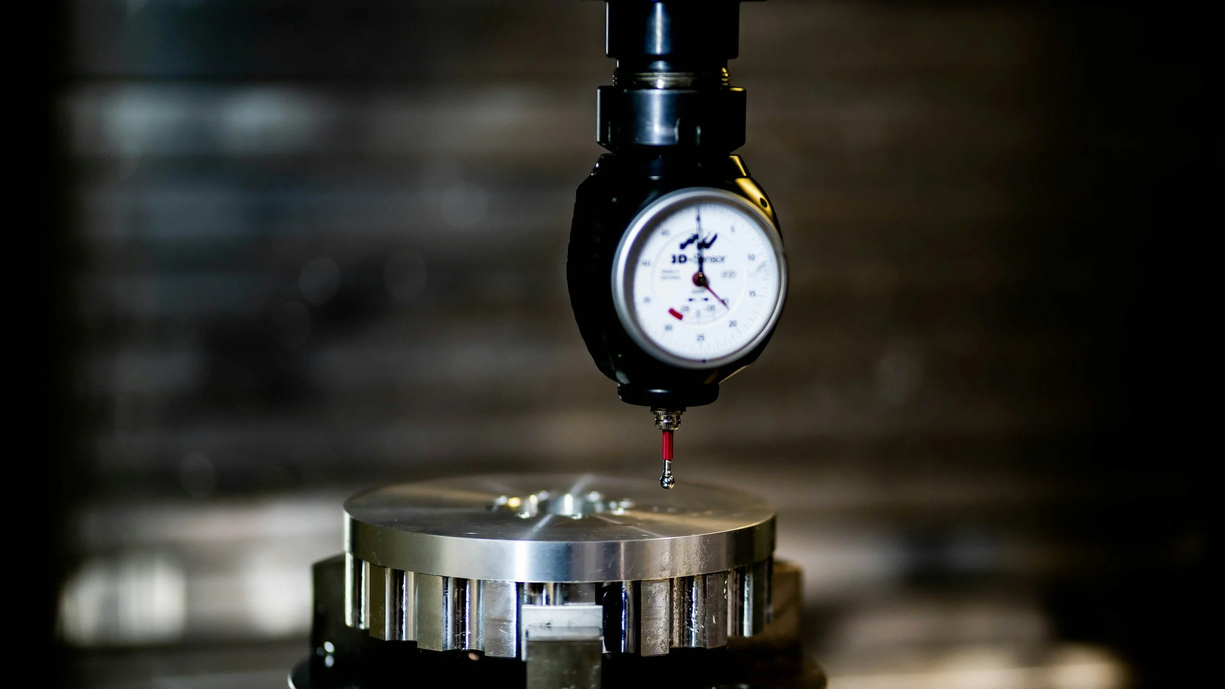 Close-up of a dial indicator measuring a metallic component during a machining process in a factory or laboratory setting.