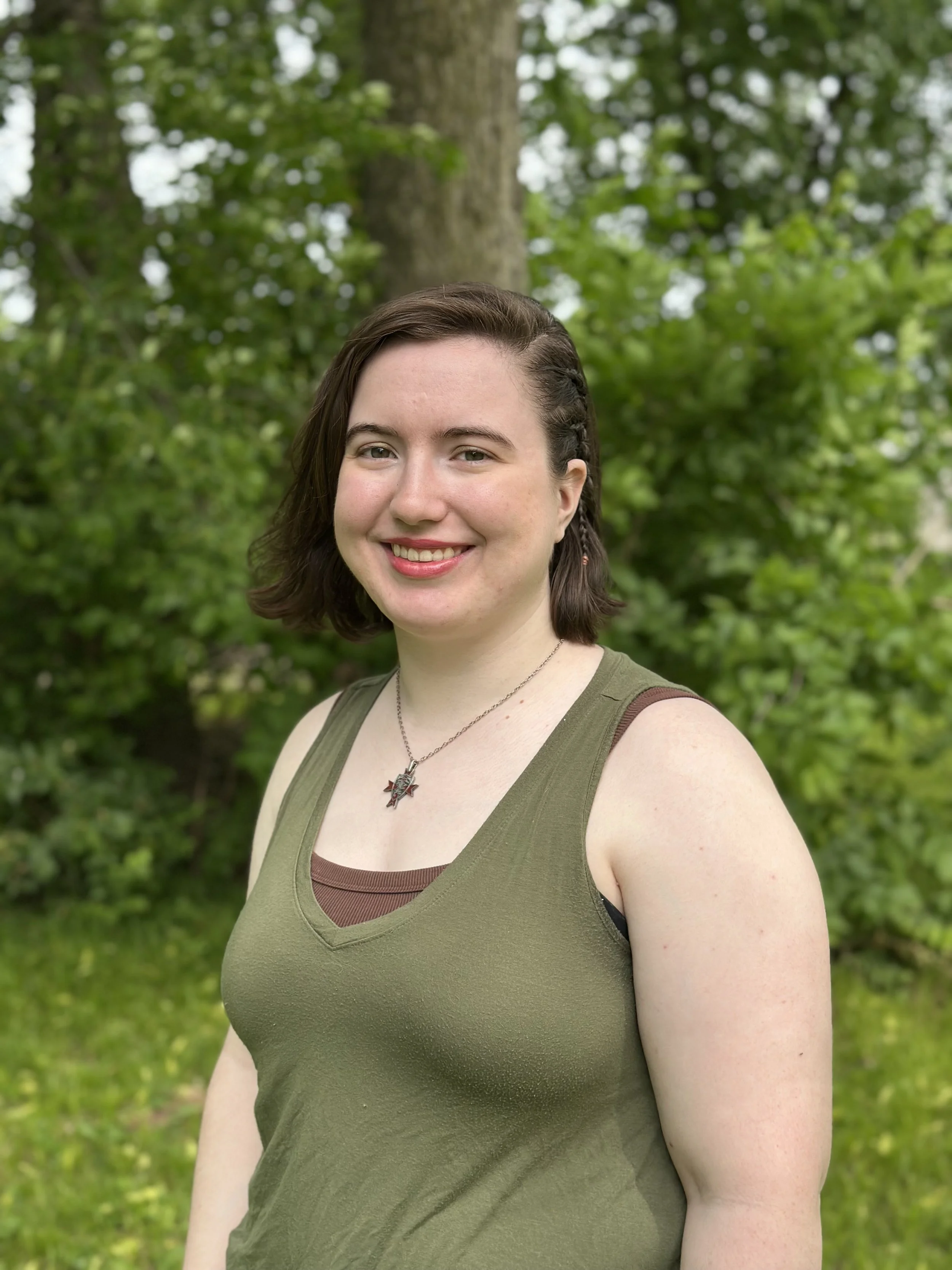 Person standing in a wooded area, wearing layered tops, surrounded by greenery.