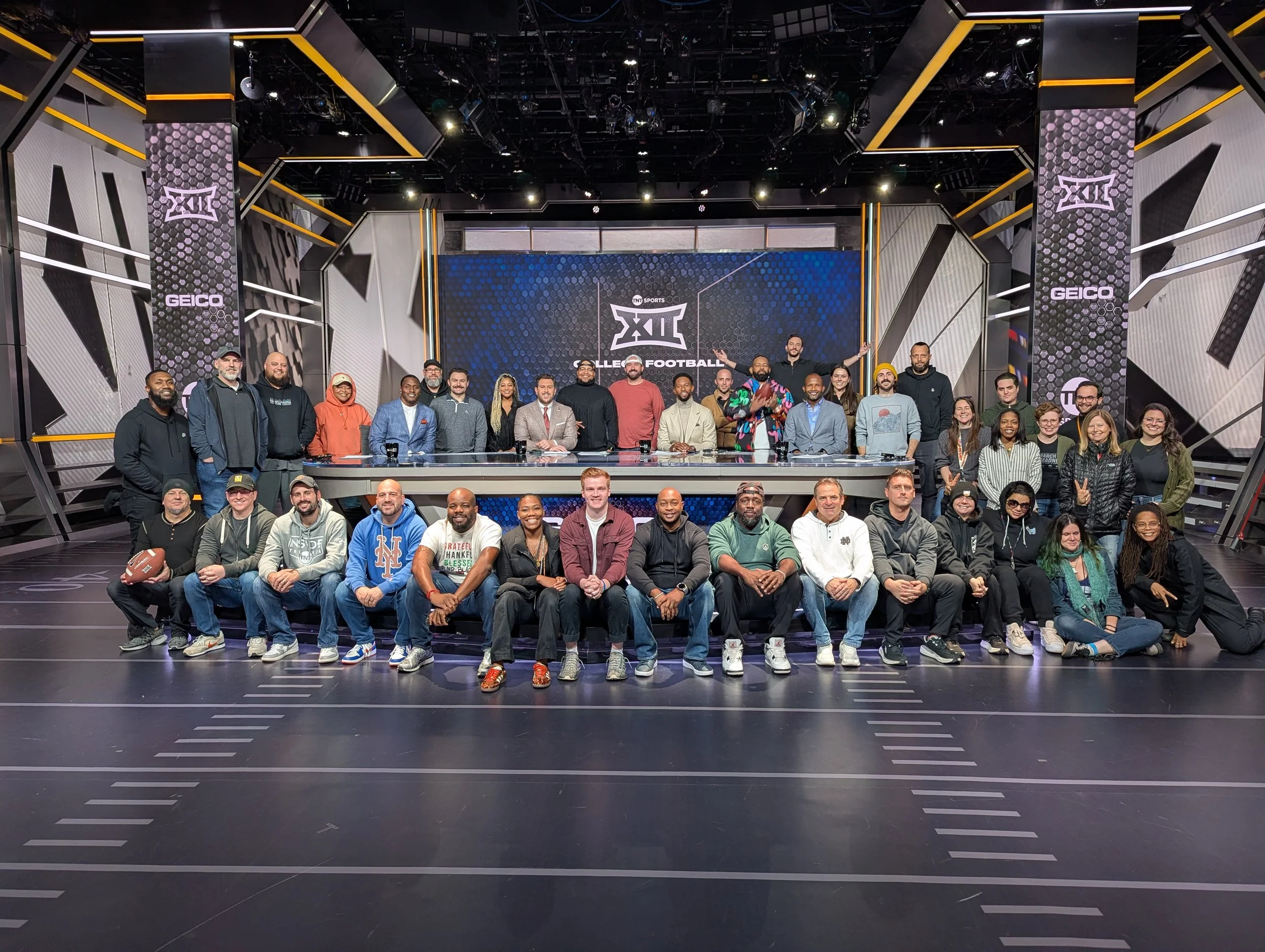 Group photo of diverse people standing and sitting on a football field with a studio set in the background featuring 'Big 12 College Football' banner and studio logos.