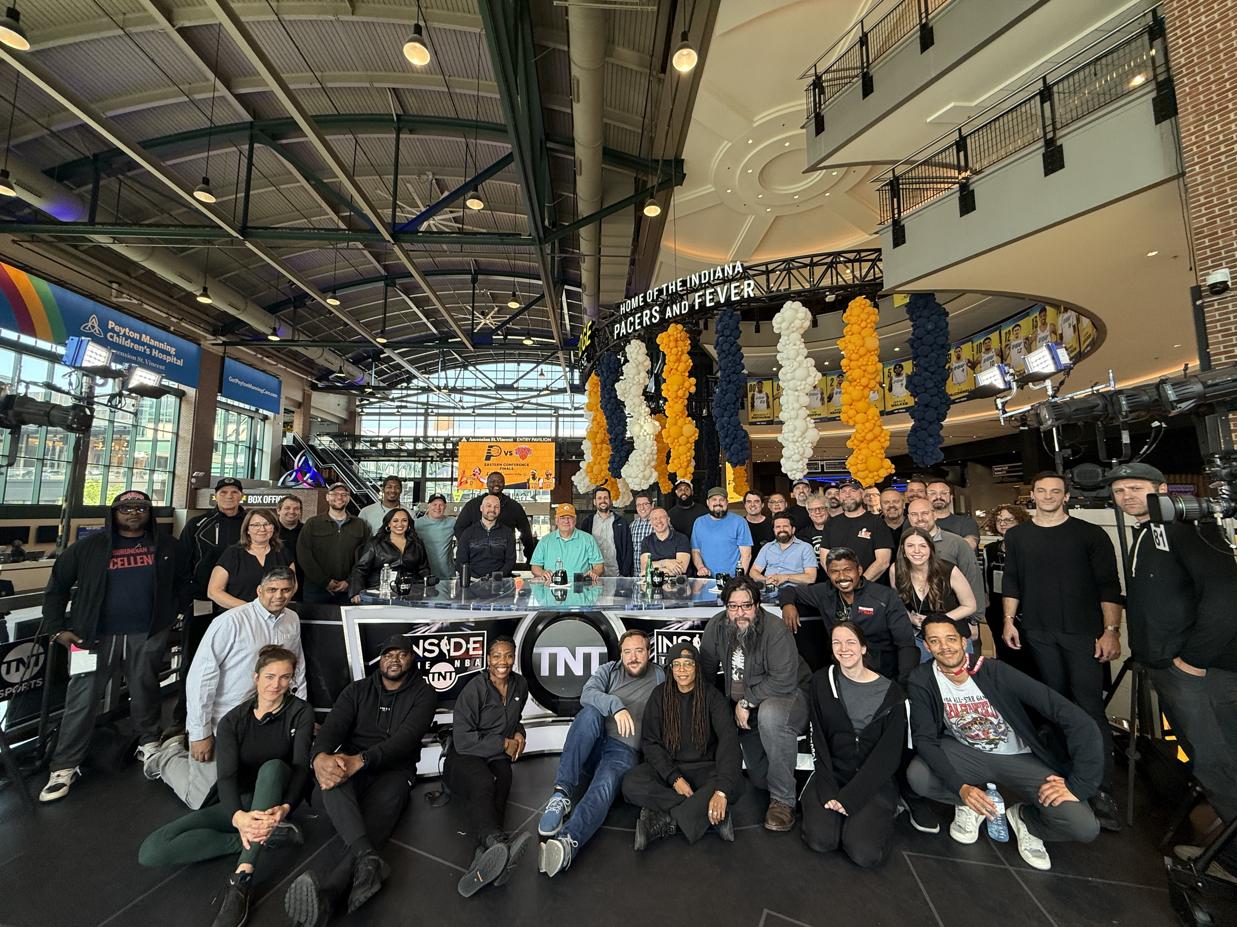 A large group of people gathered in an indoor arena with high ceilings, decorated with balloons and banners, including a sign that says "Home of the Indiana Pacers and Fever" and screens showing basketball game info. The people are smiling and posing