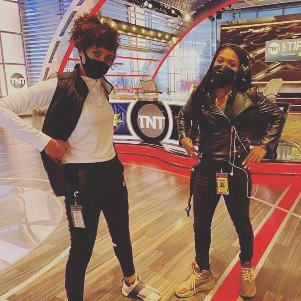 Two women wearing black face masks and casual clothing standing inside a TV studio with TNT and sports broadcasts in the background.