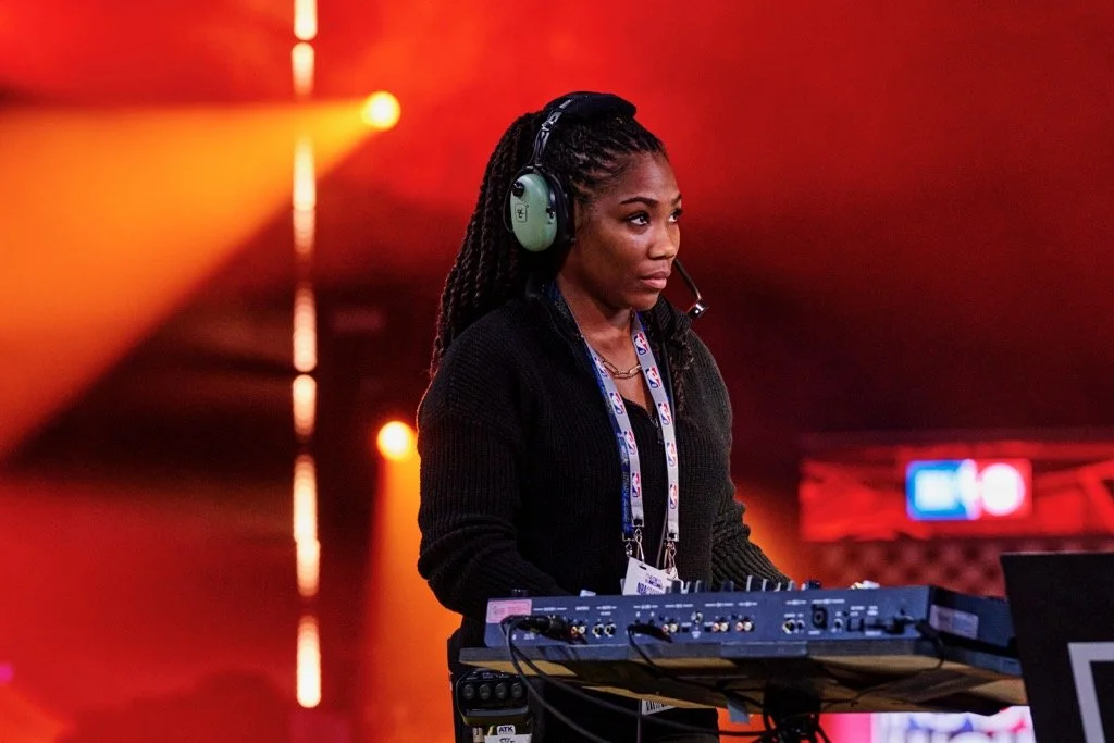 A woman DJ wearing headphones and a headset, standing behind a DJ console, with a colorful stage lighting in the background.