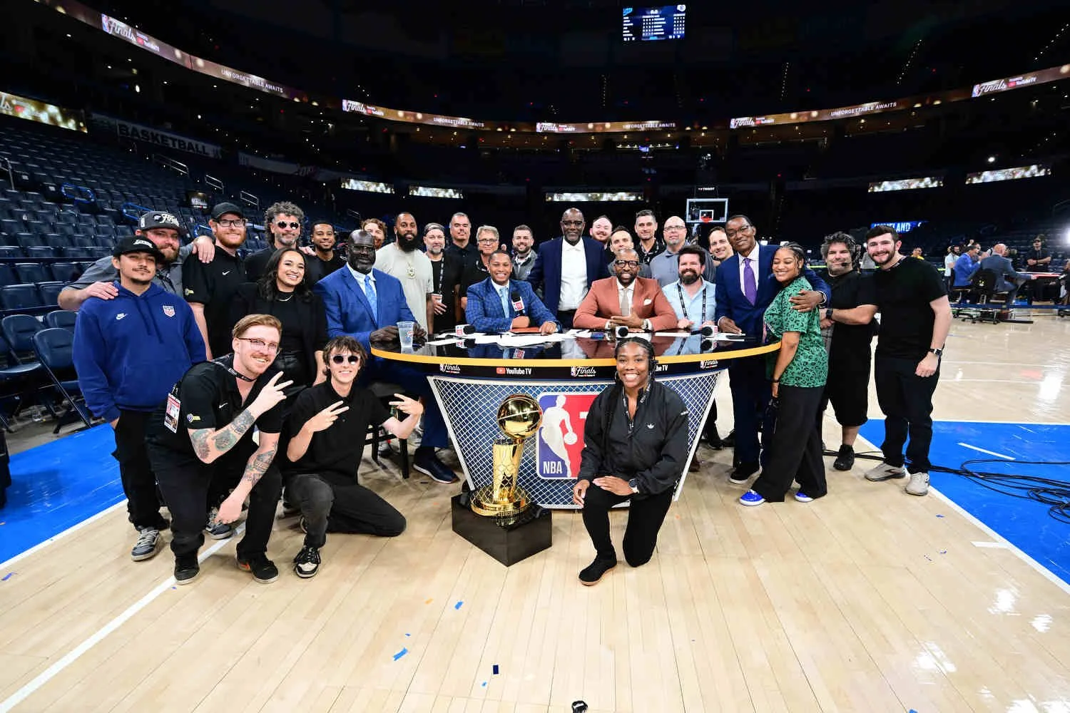 Group of people, including NBA players, coaches, and staff, celebrating on a basketball court with an NBA championship trophy, after a game or tournament.