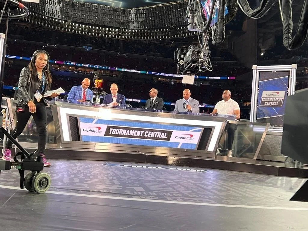 Sports broadcast studio with five men seated at a desk labeled "Tournament Central" and a woman standing with headphones, holding papers, in front of equipment and camera gear.