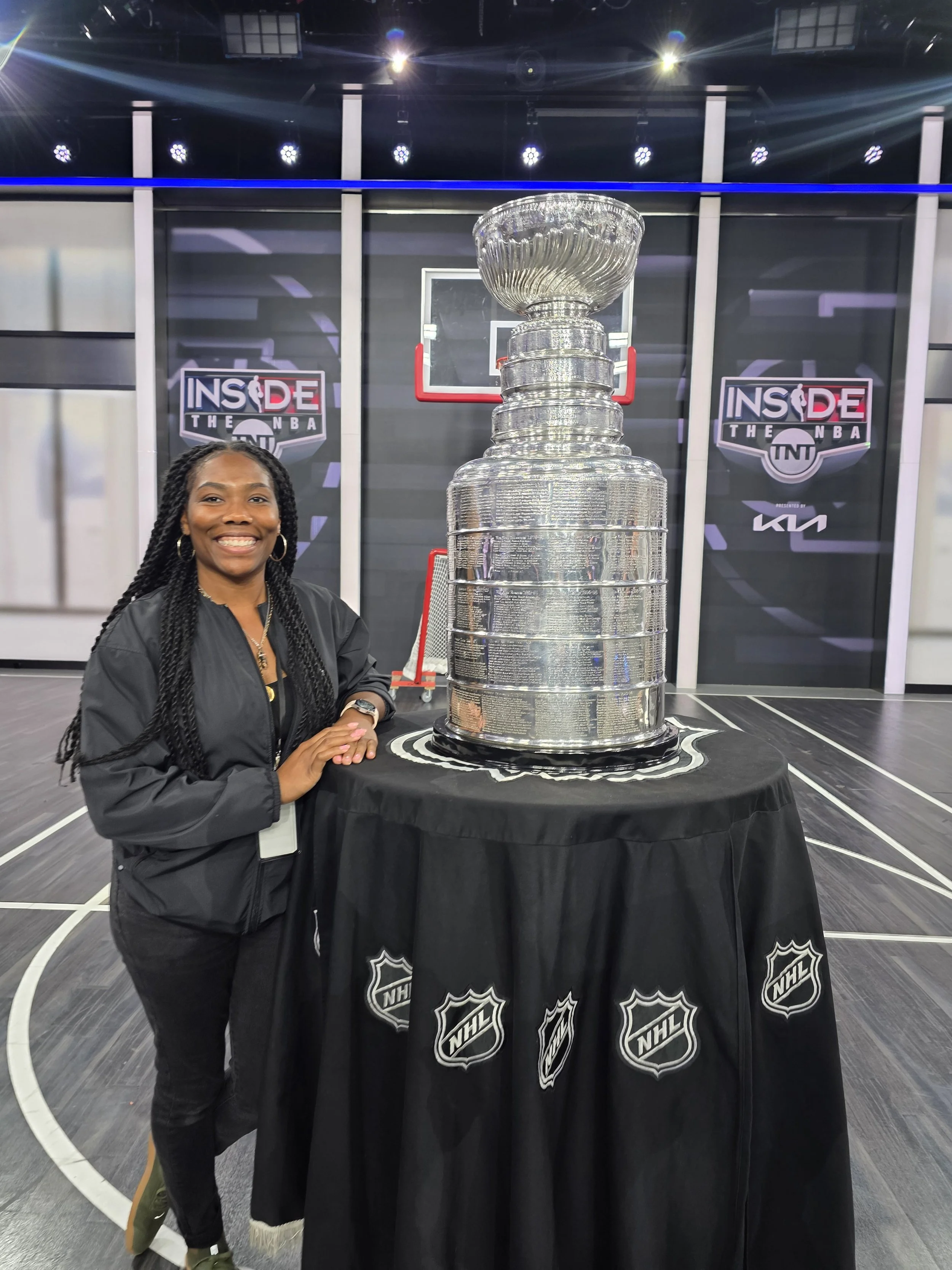 A woman with braids and hoop earrings smiling while standing next to the Stanley Cup trophy at an indoor event. The background shows logos for 'Inside the NBA,' TNT, KIA, and NHL.