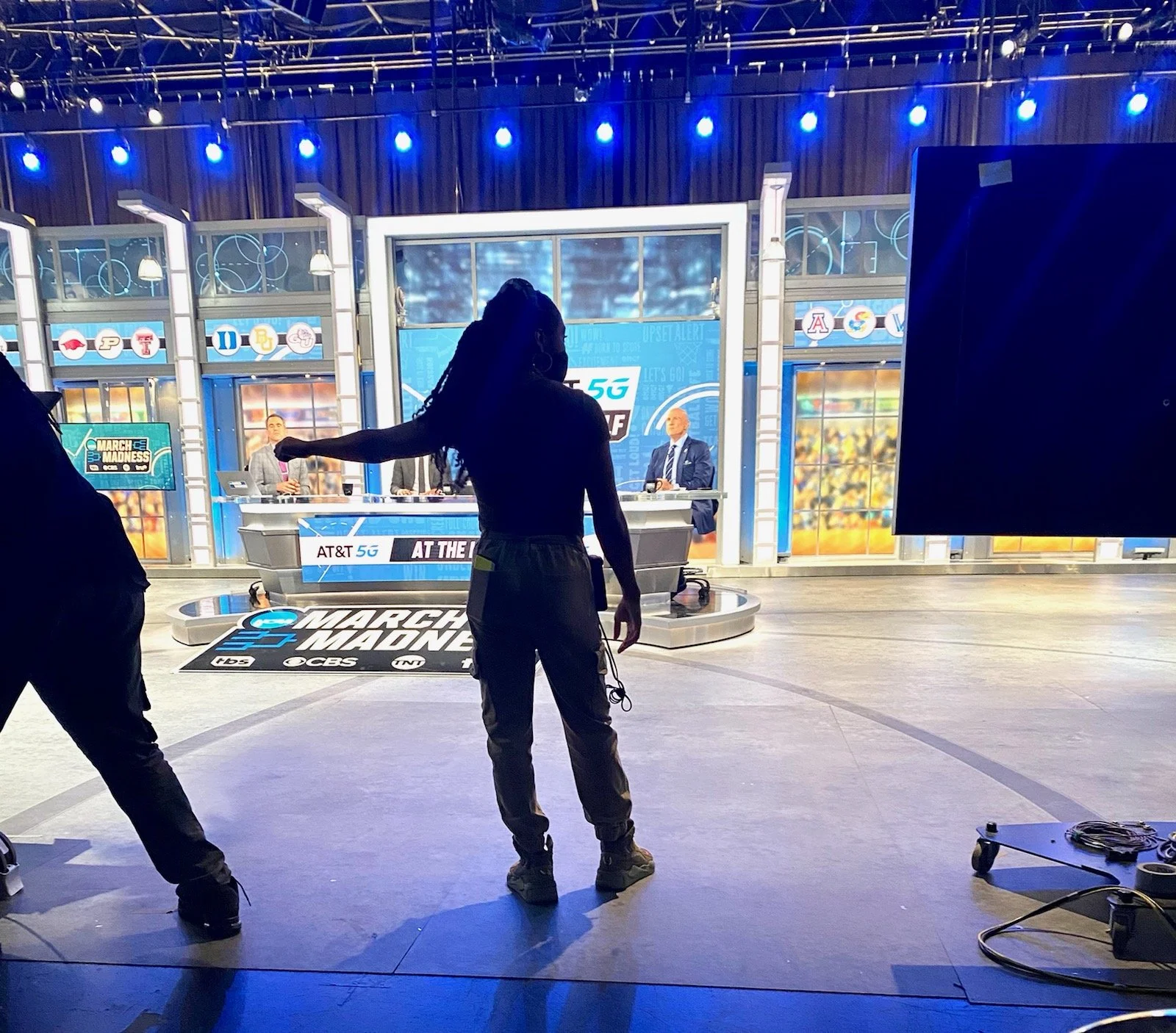 Behind the scenes of a televised sports broadcast, a person with dreadlocks is standing in front of a news desk, with three analysts seated at the desk. The studio is brightly lit with blue lighting, and the floor has the March Madness logo. Bright s