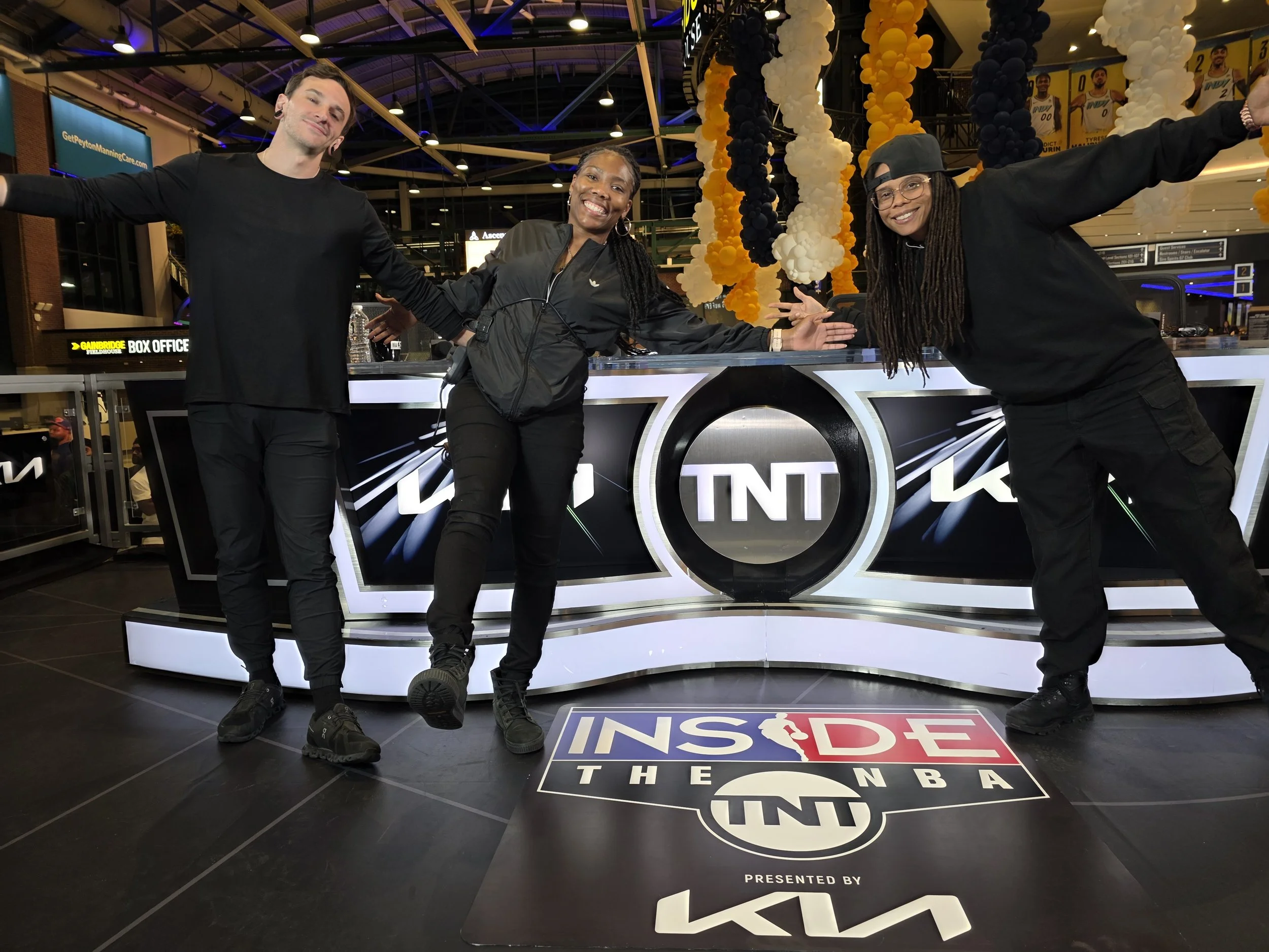 Three people standing in front of a TNT NBA logo at an indoor event, smiling and posing with their arms spread out.