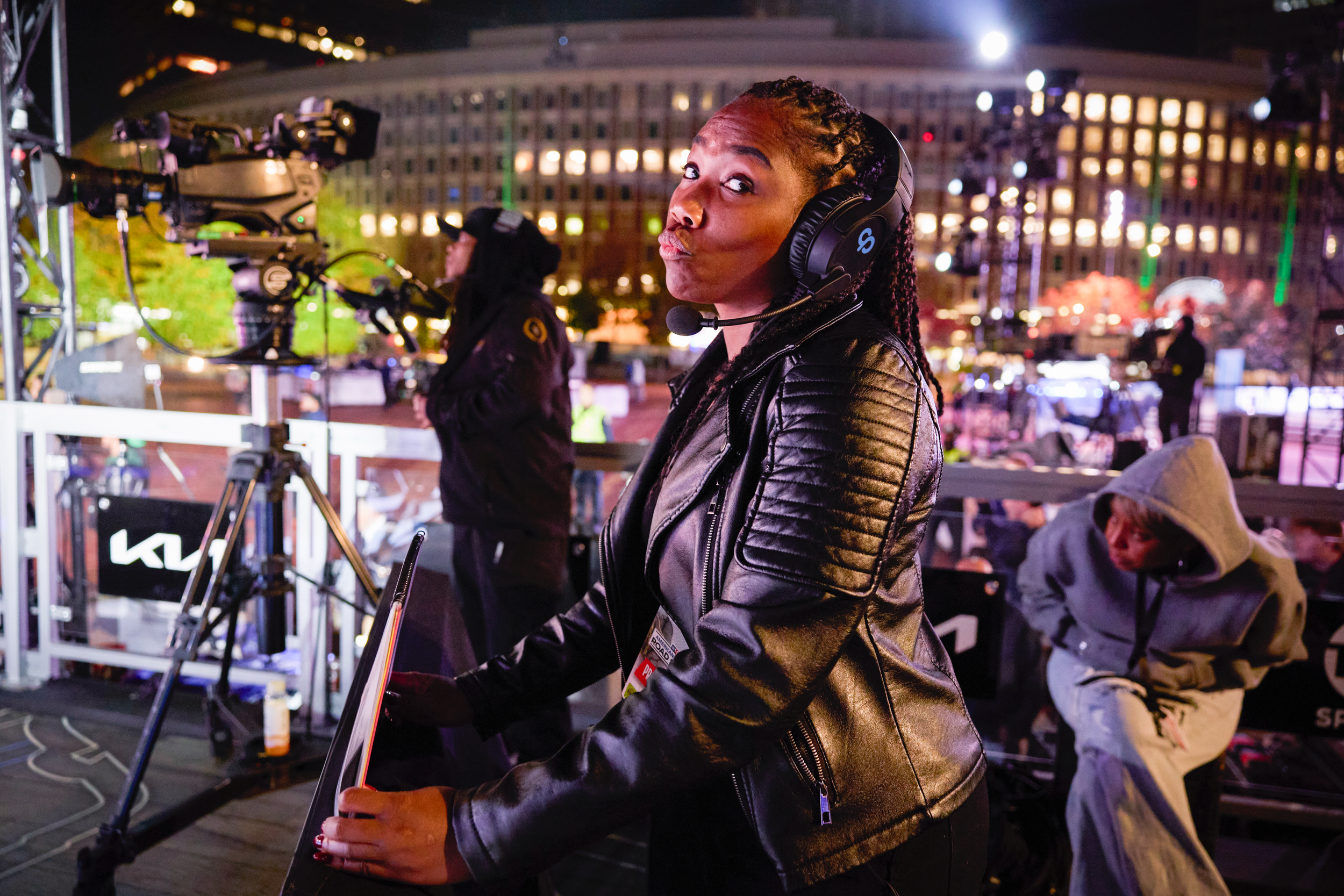 A woman wearing a black leather jacket and large headphones is at an outdoor event at night. She appears to be preparing to speak or perform, with a camera crew and other people in the background. City buildings with lit windows are visible behind he
