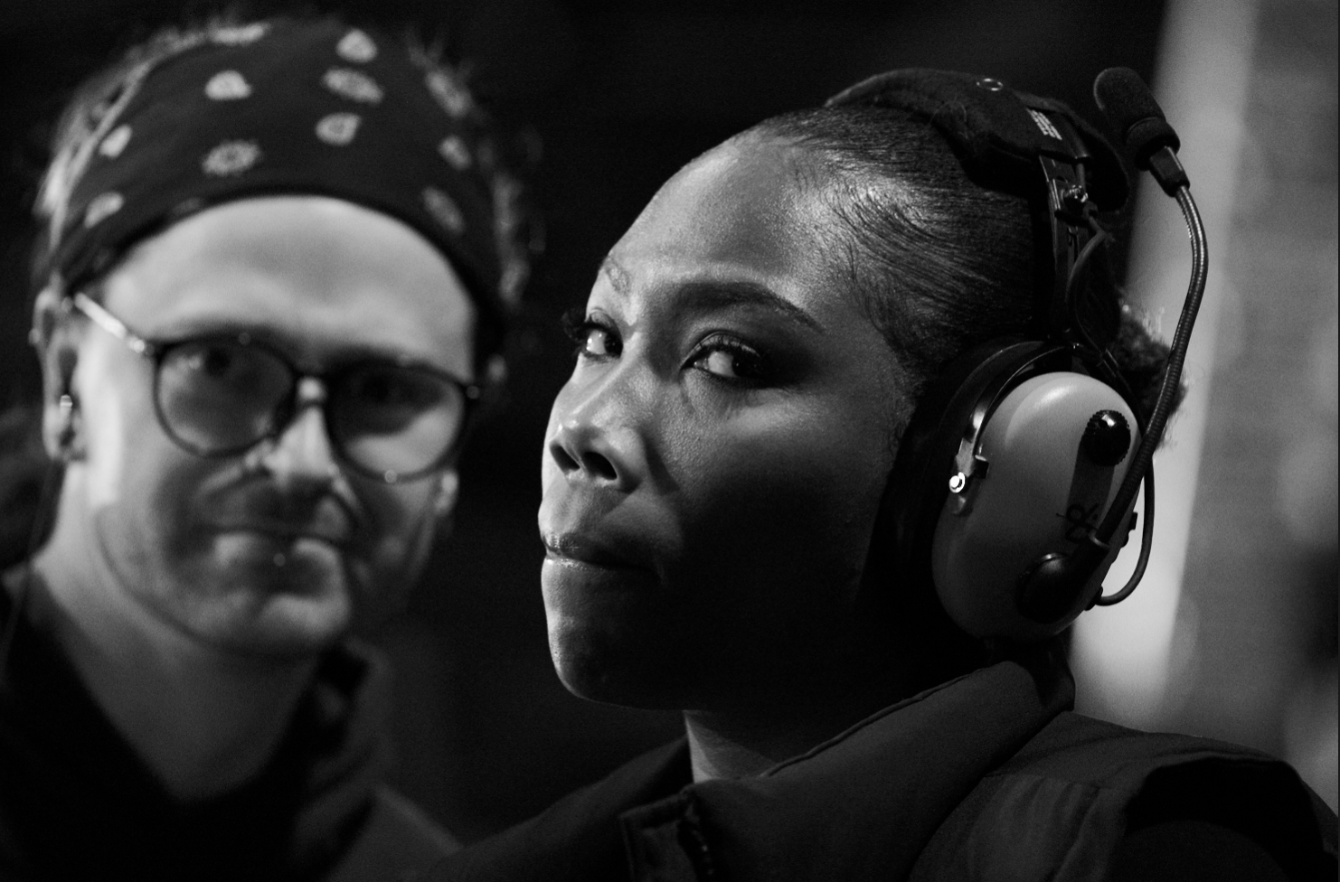 A close-up black-and-white photo of a woman wearing large headphones, with a serious expression, and a man with glasses and a headscarf in the background.