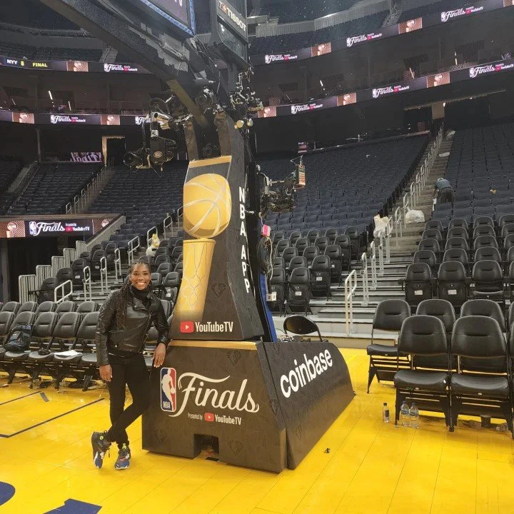 A woman smiling and standing next to a basketball hoop with a NBA Finals and Coinbase logo on it, on a basketball court with empty seats in the background.