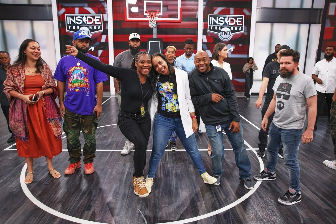 A group of diverse people standing on a basketball court, smiling and posing for a photo, with a basketball hoop and two screens displaying the 'Inside the NBA' logo in the background.