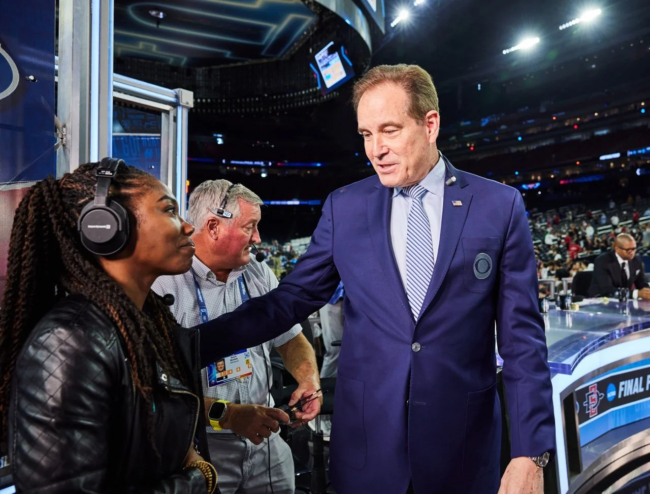 An interviewer and a man in a suit having a conversation at a sports event, with a large audience in the background.