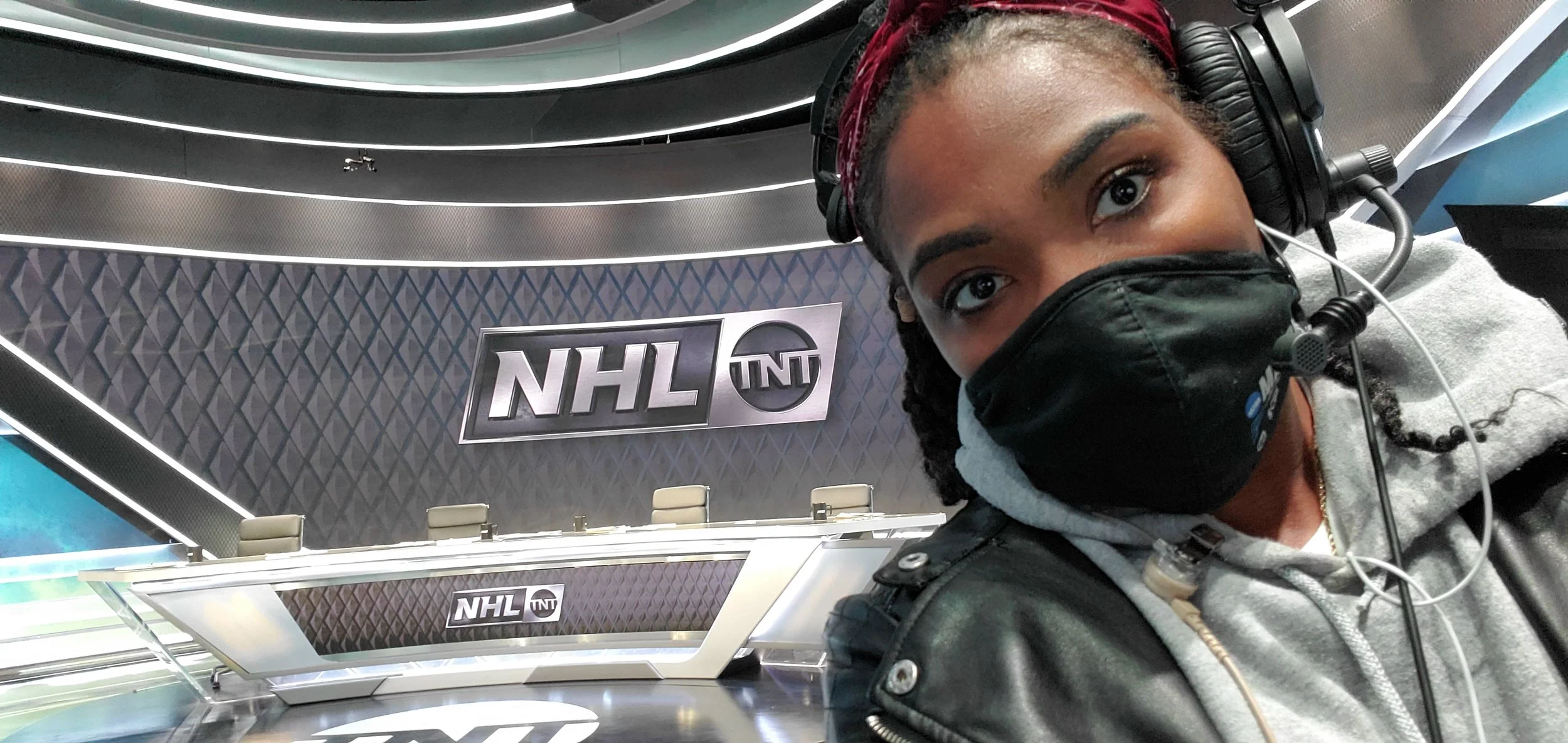 A woman wearing a black face mask and headset taking a selfie in a broadcast studio with NHL and TNT logos on the background.