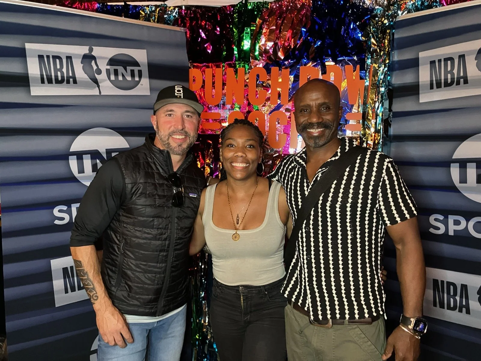 Three people standing in front of a backdrop with the NBA, TNT, and Sportsnet logos, and colorful tinsel decoration. The backdrop also has bright pink text that reads "PUNCH A COACH." All three are smiling.