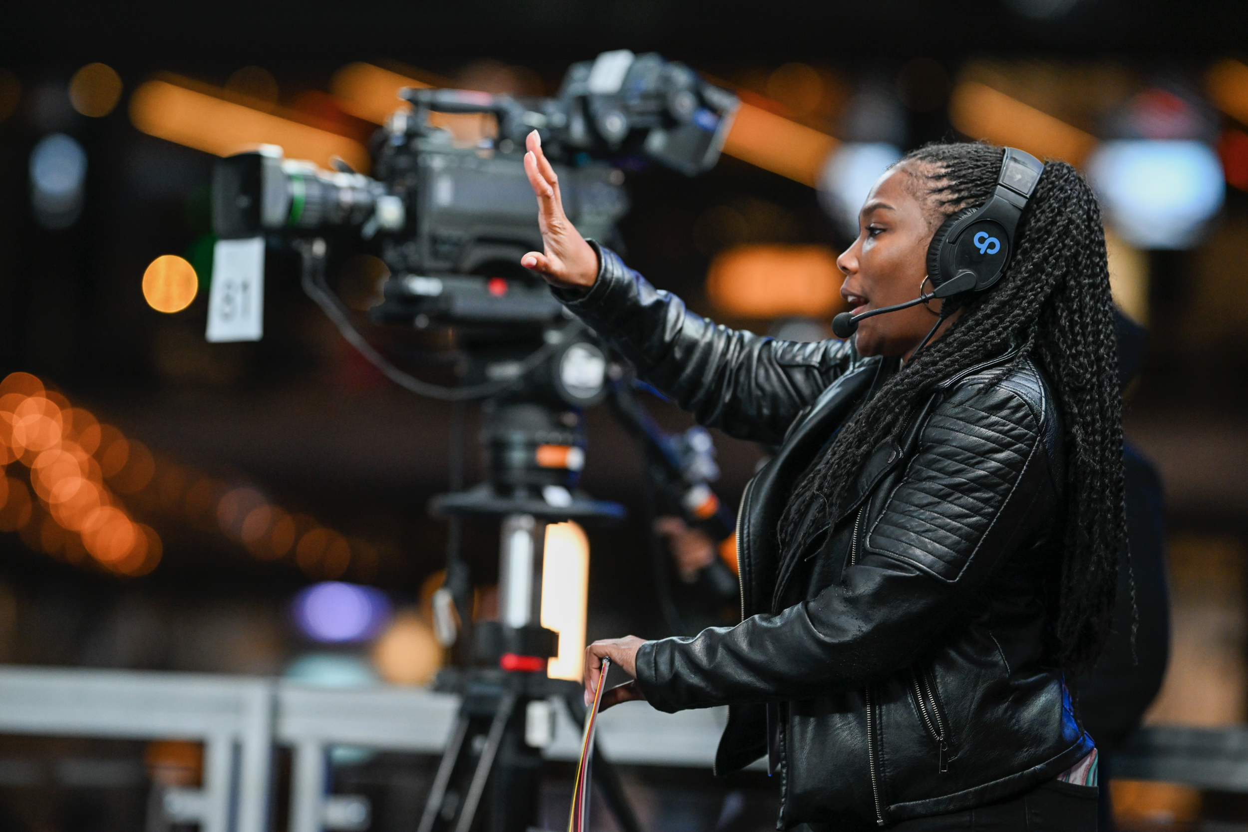 A woman with braided hair, wearing a leather jacket and headphones, is adjusting a professional video camera on a tripod during an outdoor event at night.