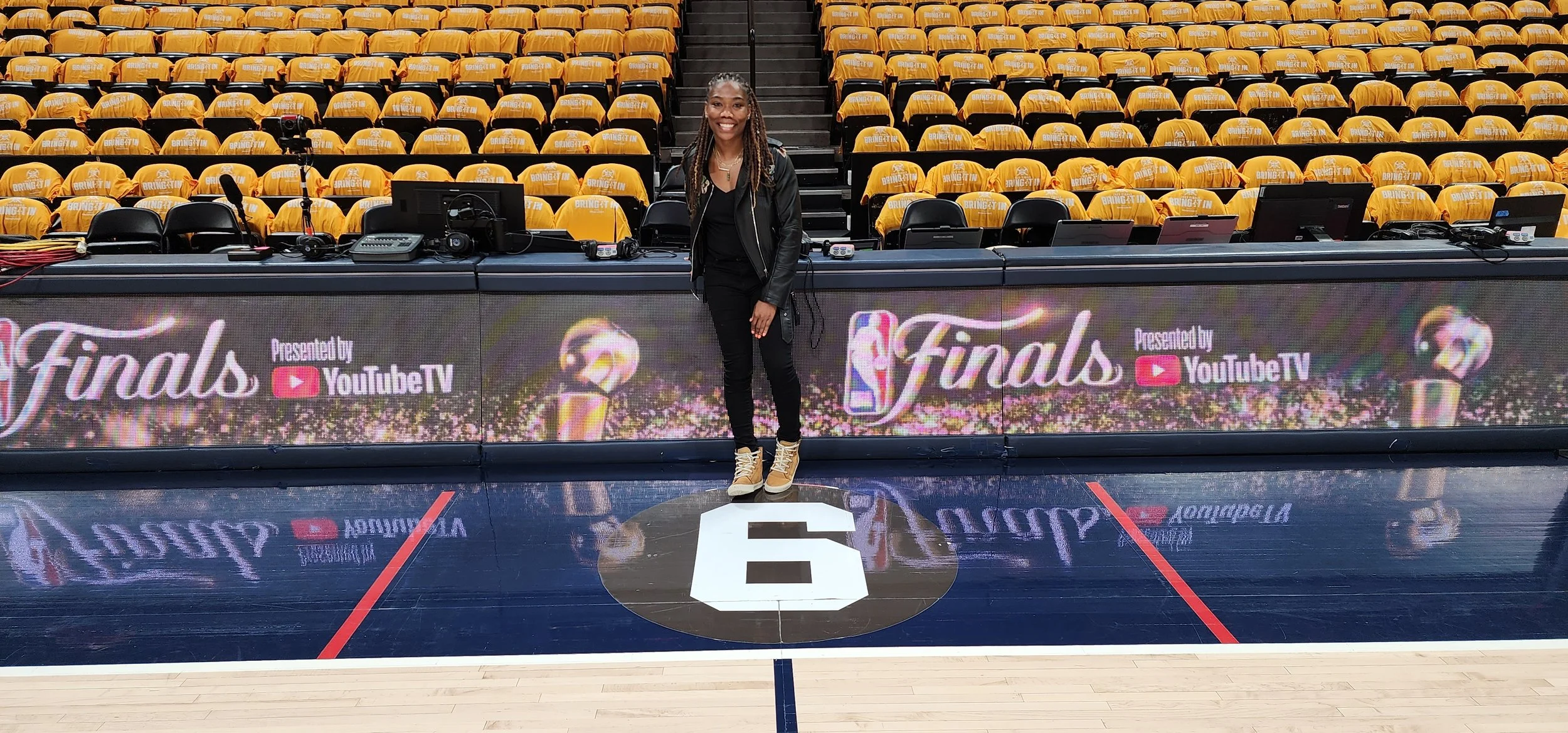 A woman standing on a sports court in front of a large digital banner with the words 'Finals Presented by YouTube TV' and a red play button logo. She is smiling, wearing black clothing and tan boots, with yellow seats and some audio equipment behind 