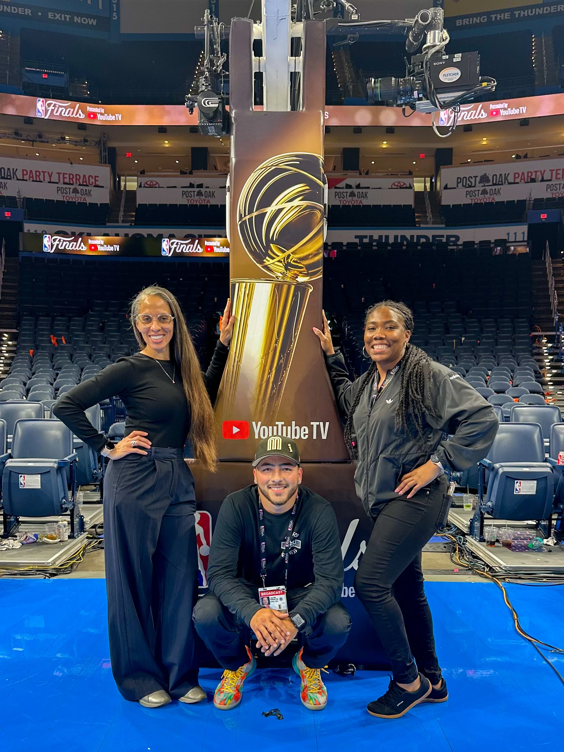 Four people pose with an NBA championship trophy on a basketball court. Two women stand on either side, one man kneels in front holding a broadcast badge, and another person is seated, all smiling. The background shows empty arena seats and visible d