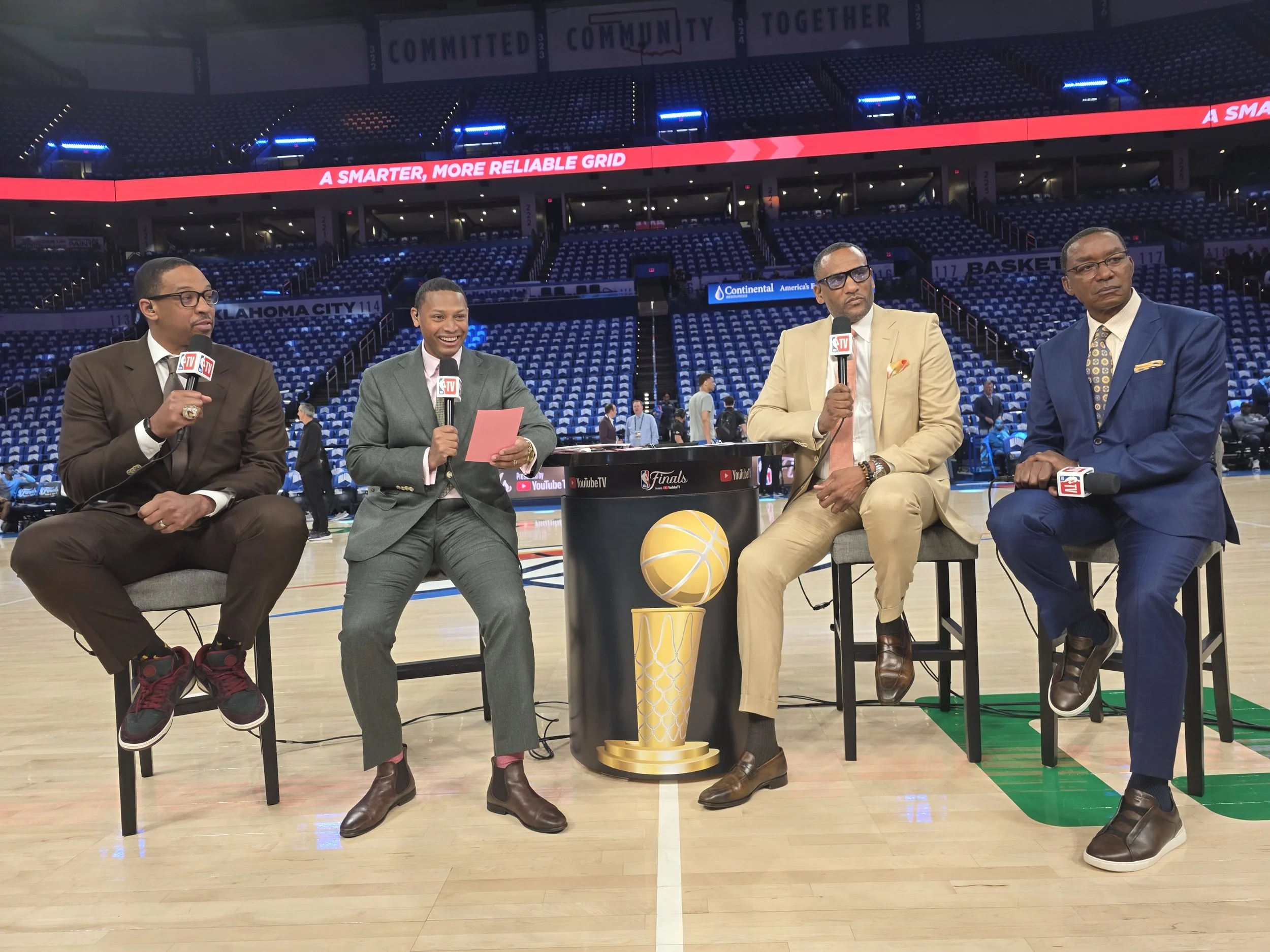Four men dressed in suits sitting in a row on chairs in a basketball arena, holding microphones with NBA logos, with a basketball and a trophy in front of them. The arena is mostly empty with a few people in the background and signs overhead, includi