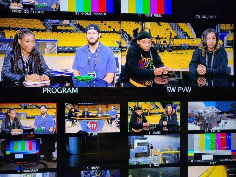 Four individuals sitting at a news desk in a sports arena, with a large TV monitor above displaying test patterns and the words "PROGAM" and "SW PVW." The scene appears to be a behind-the-scenes view of a sports broadcast production.