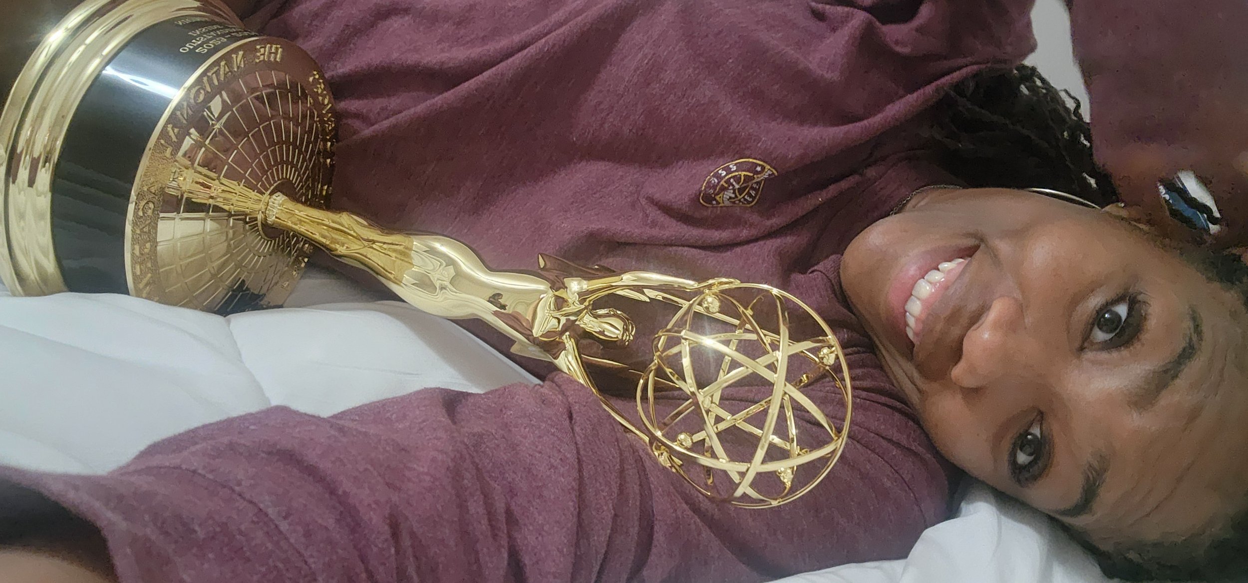 A woman lying in bed smiling with Emmys on her chest, wearing a maroon sweatshirt.