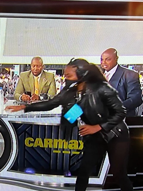 A woman in black pushing a shopping cart or container away from a television studio set with two men standing behind her, one in a brown jacket and the other in a suit, with a large monitor and the words 'CARMAY' visible.