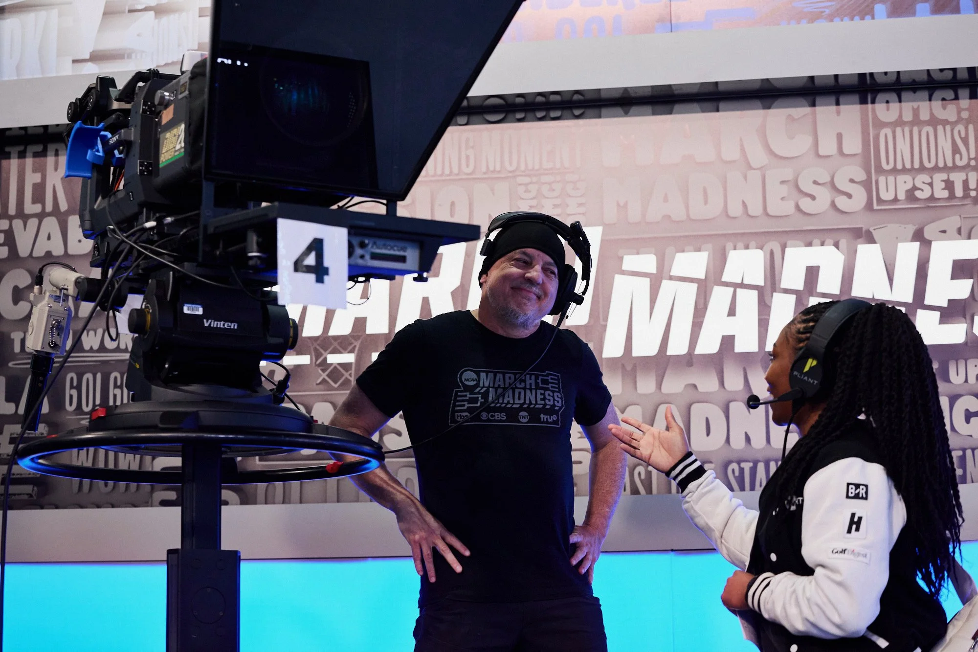 Man wearing March Madness t-shirt and headphones smiling and talking to a woman with headphones, in front of camera equipment and a poster with words related to March Madness.